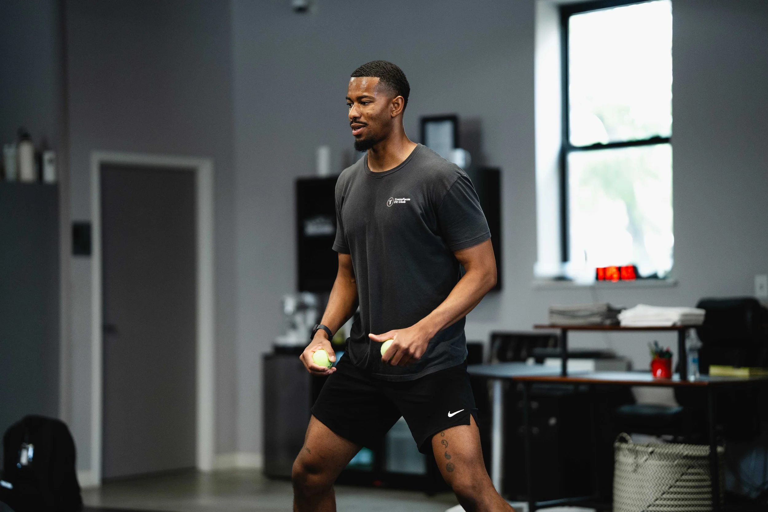 A man in a black athletic t-shirt and shorts holding tennis balls in a gym.