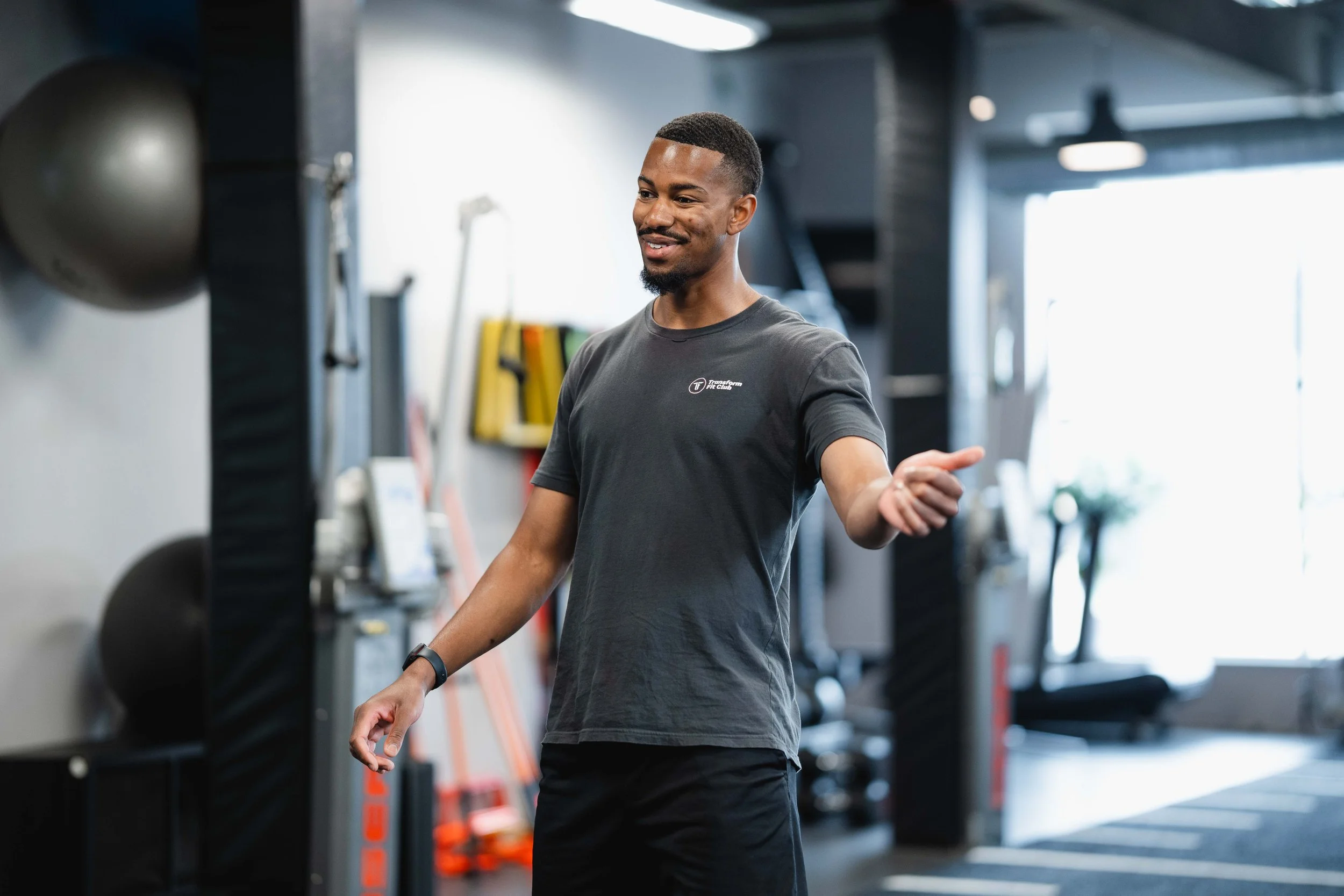 A man standing in a gym, smiling, wearing a gray t-shirt with a logo, and looking down at his hand.