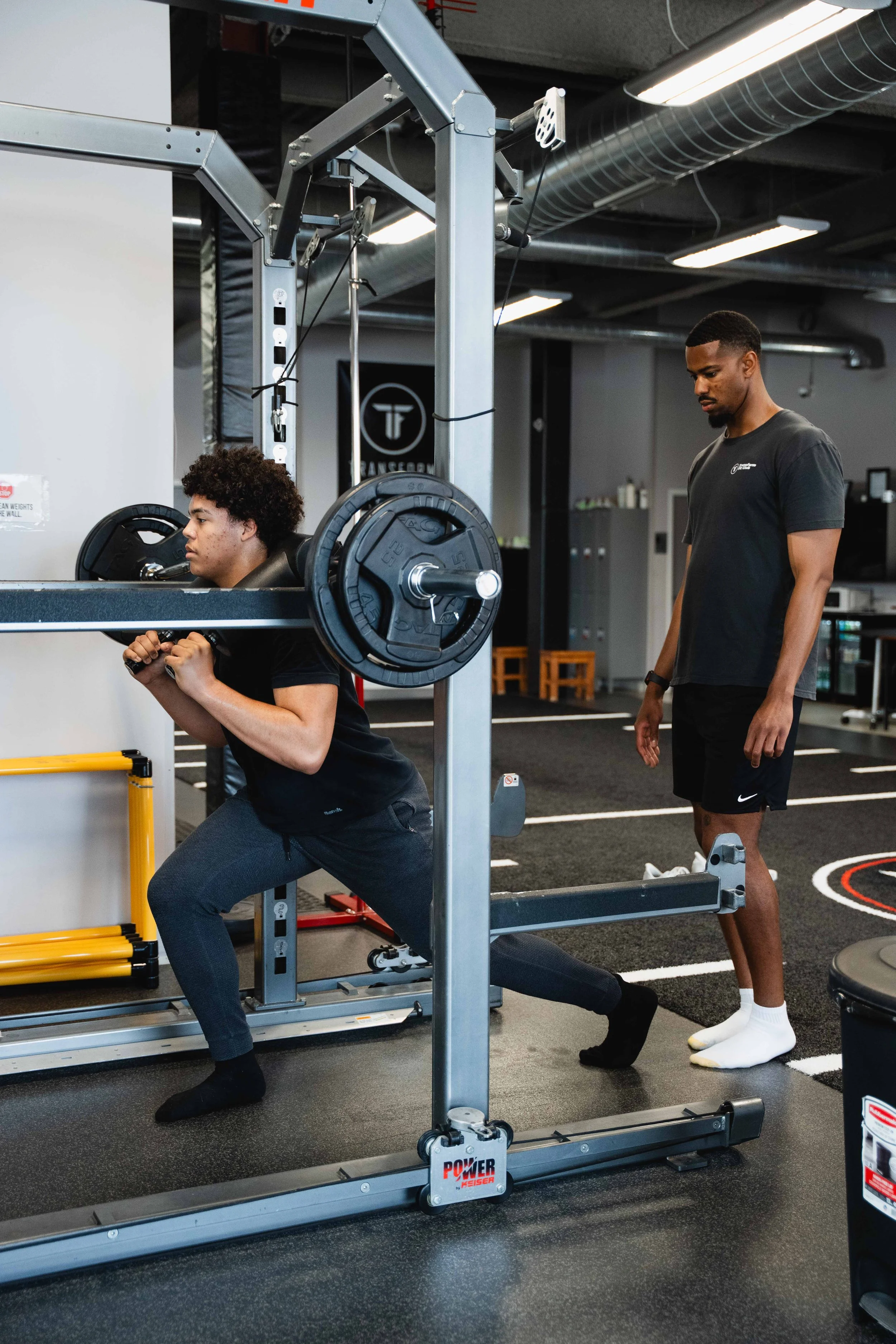 A person doing a squat exercise with a barbell loaded with weights on their shoulders, while a trainer stands nearby in a gym.