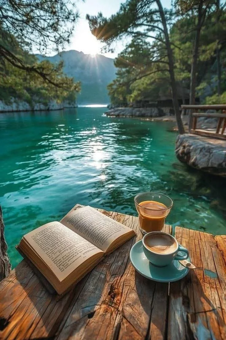 Ein Buch, ein Glas Tee und eine Tasse Kaffee auf einem Holzbrett am Ufer eines klaren, grünen Sees mit Bäumen und Bergen im Hintergrund, Sonnenlicht spiegelt sich im Wasser.