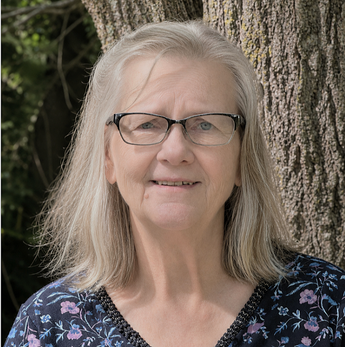 An woman with light gray hair, glasses, and a floral top standing outdoors in front of a tree.