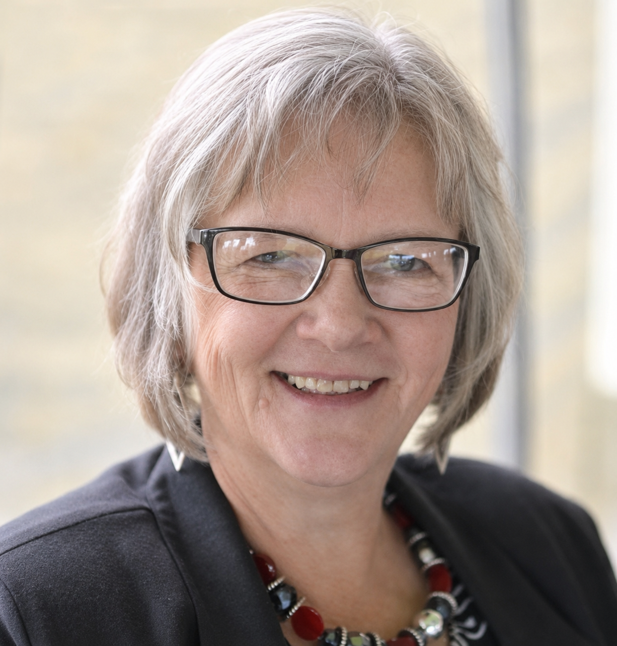 A smiling woman with gray hair and glasses, wearing a black blazer and a red and black beaded necklace, in front of a bright window.