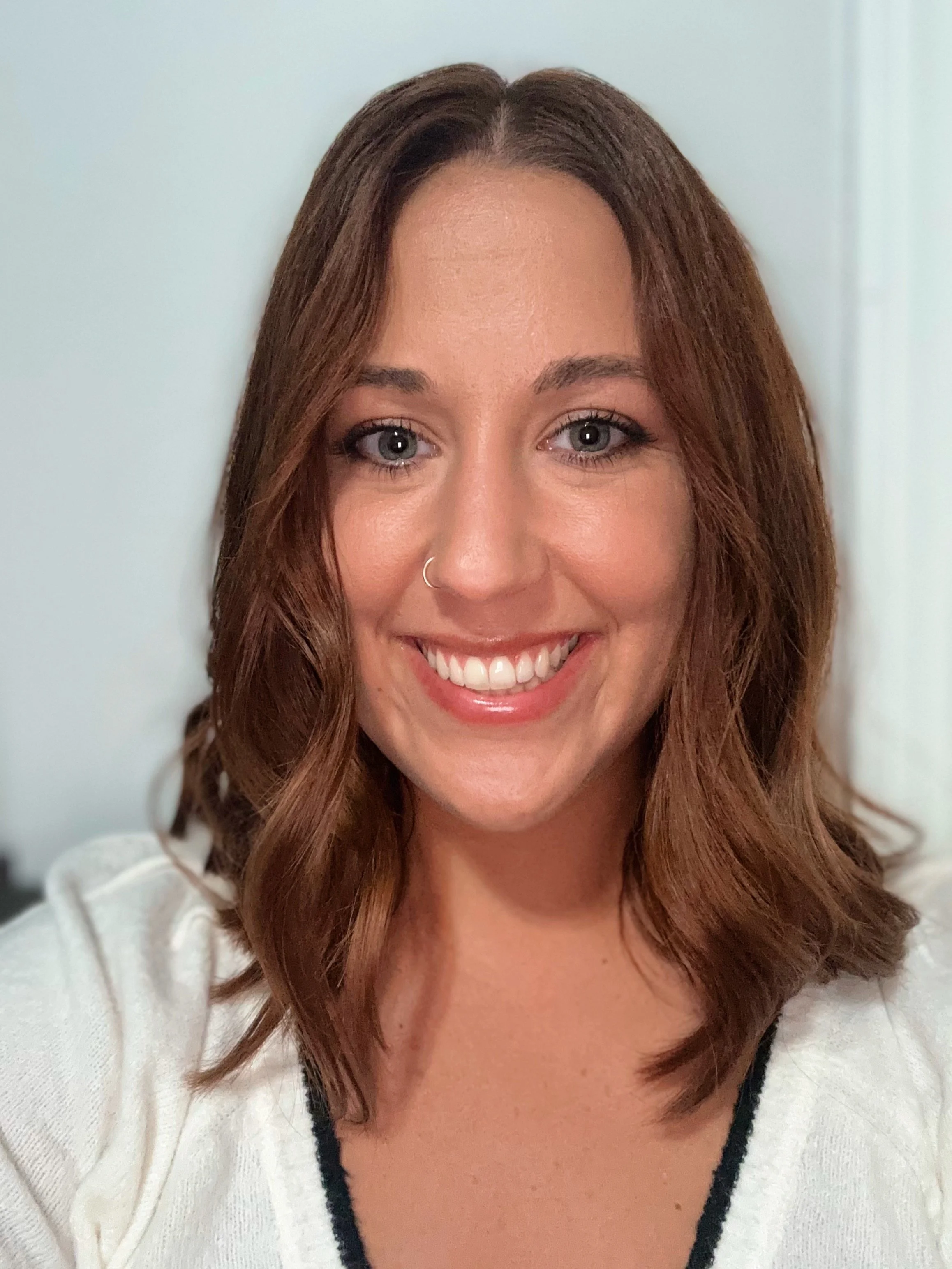 Close-up photo of a smiling woman with light blue eyes, brown wavy hair, a nose ring, and wearing a white top with black trim.