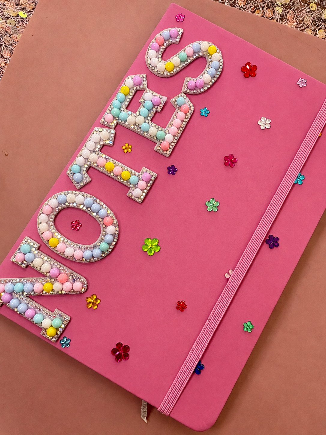 Pink notebook with the word 'LOVE' written in large, decorative letters made of pastel-colored beads and pearls, surrounded by small, colorful flower-shaped sequins.