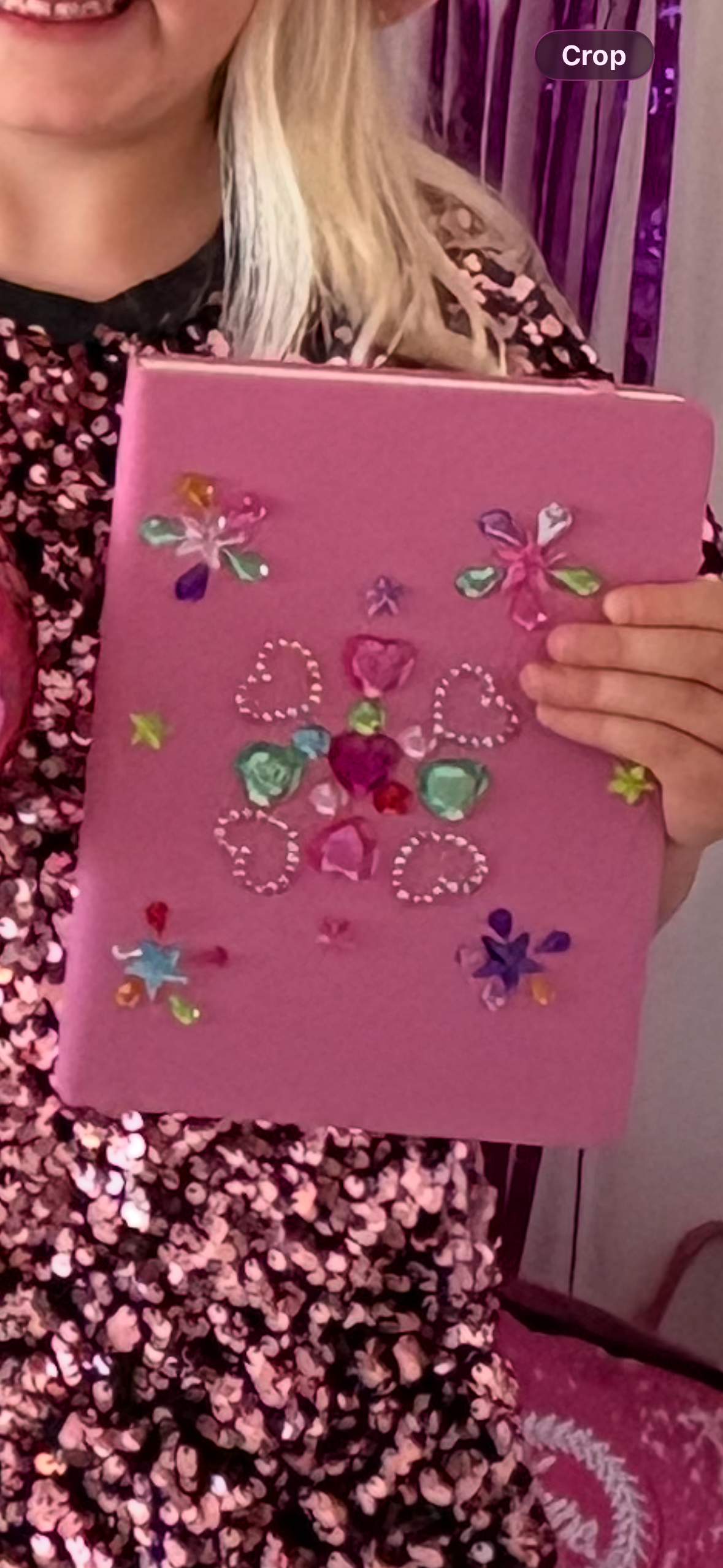 A person wearing a floral dress is holding a pink decorated notebook or scrapbook with colorful jewel embellishments and heart-shaped designs.