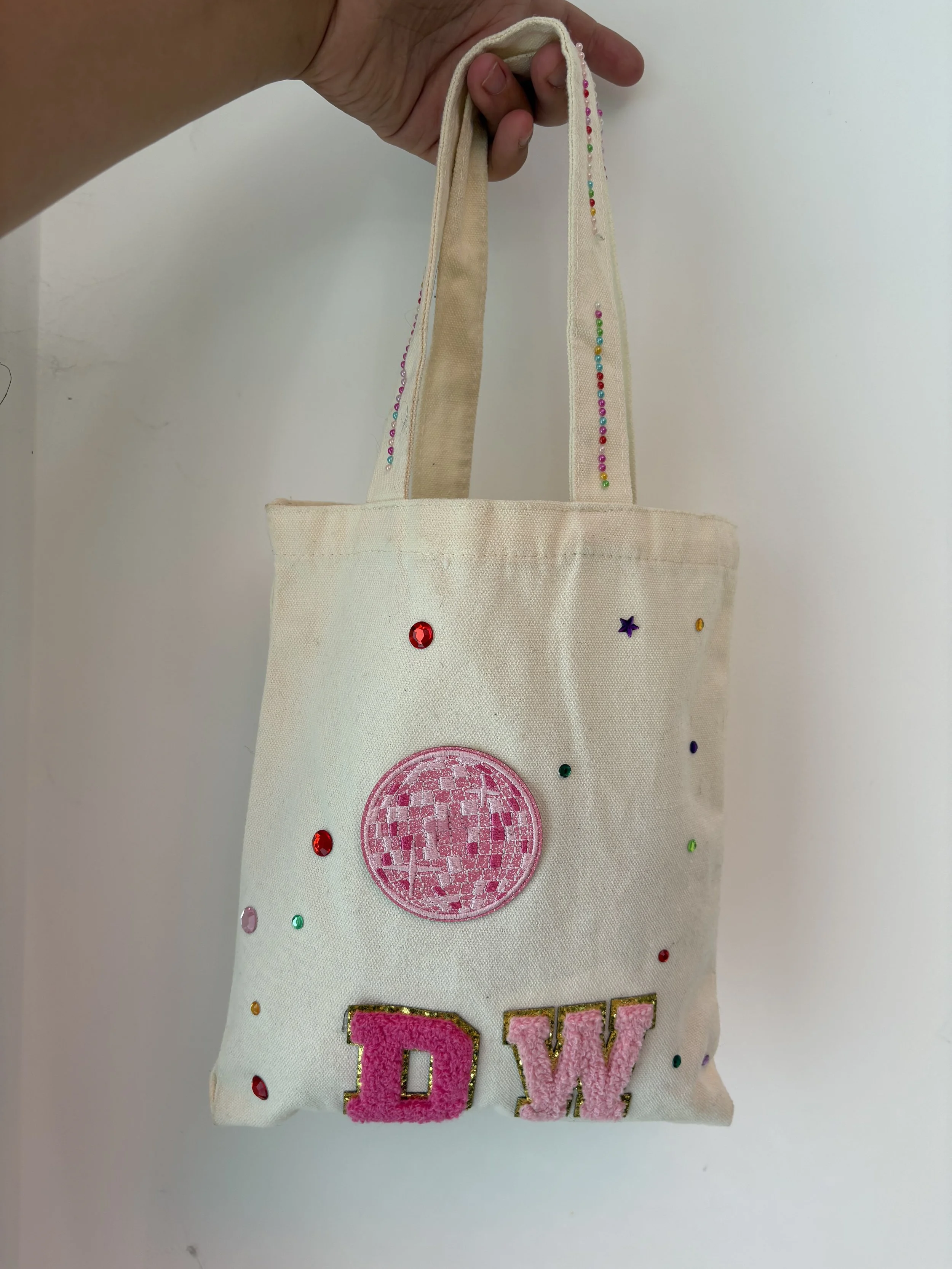 A white tote bag decorated with multicolored rhinestones, a pink embroidered circular patch, and pink and gold lettering that spells 'MOM'.
