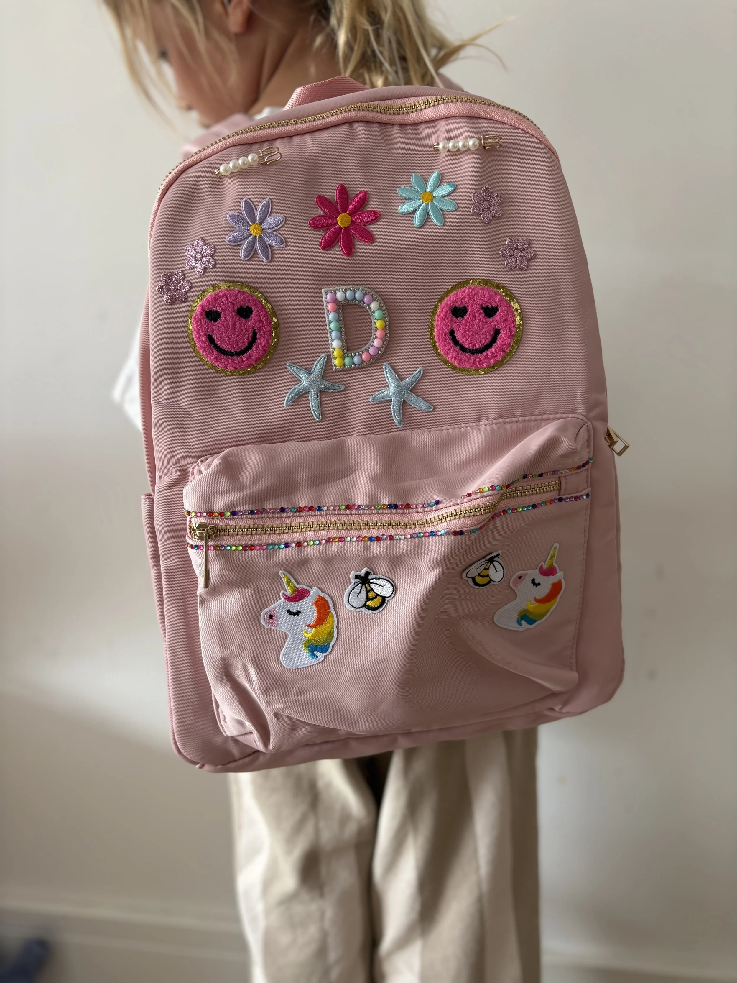 A person is holding a pink backpack decorated with embroidered and beaded patches, including smiley-faced pink circles, flowers, a letter D, starfish, and unicorn patches. The backpack has a front zipper pocket and pearl embellishments on the top.