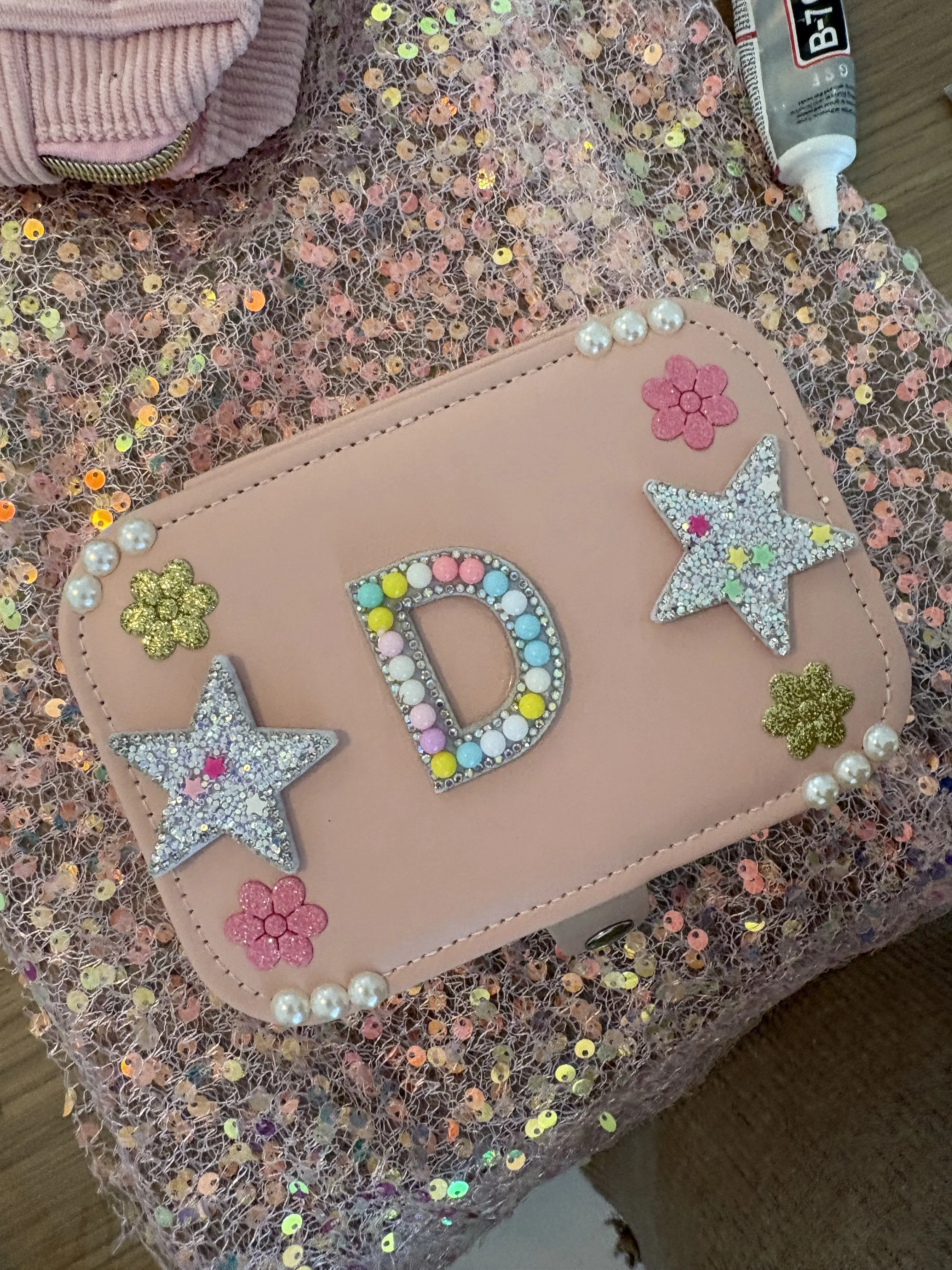 A close-up of a pink purse decorated with glittery stars, hearts, flowers, and a large letter D, with pearls along the edge. The purse is placed on a fabric with multicolored sequins and a tube of ointment or cream nearby, and a pink fabric item part