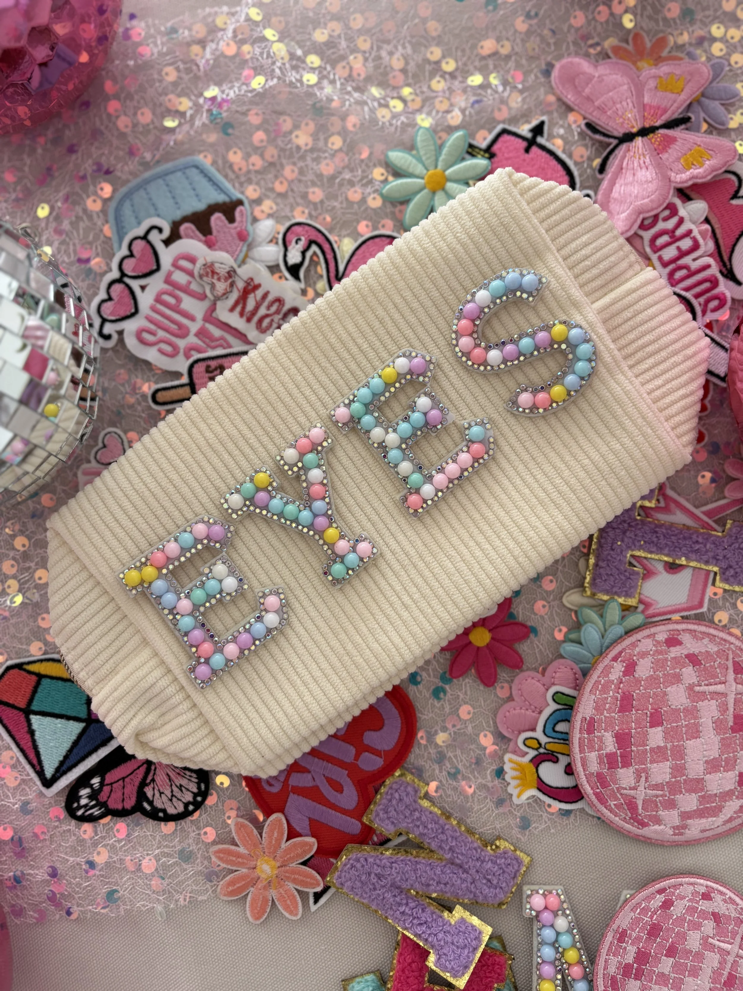 Close-up of a beige knitted accessory with colorful beaded letters spelling out 'LOVE', surrounded by various embroidered patches of flowers, butterfly, flamingo, and other designs on a pink fabric background, with sequin and glitter embellishments.