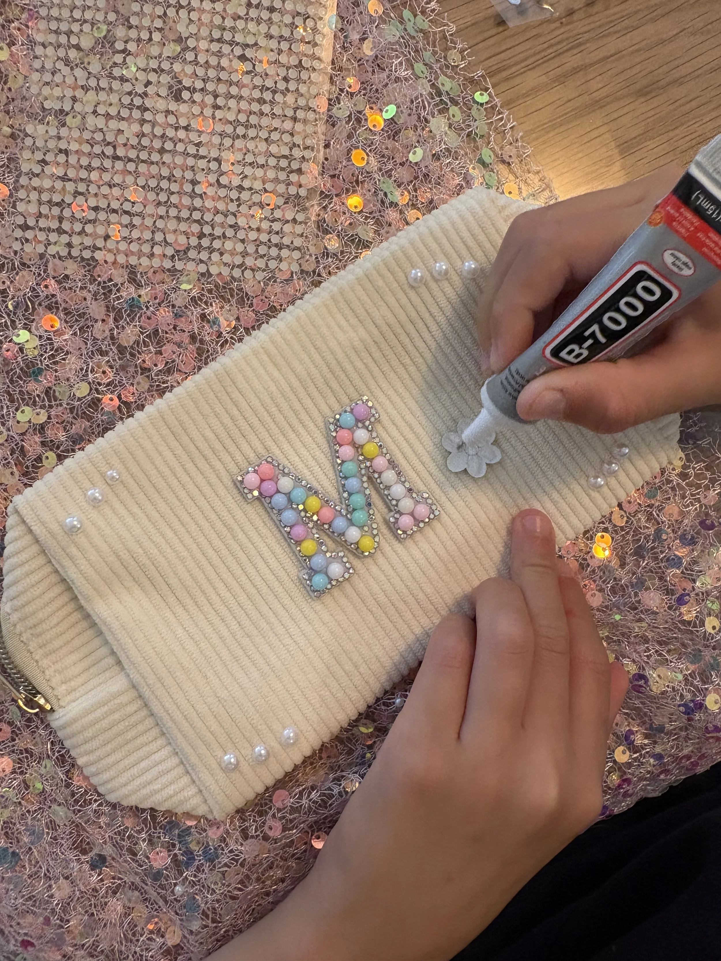 A person using a white Crayola B-7000 glue pen to attach small flower-shaped embellishments onto a beige, velvety fabric with colorful, beaded butterfly applique. The background features shiny, multicolored sequins and tinsel.