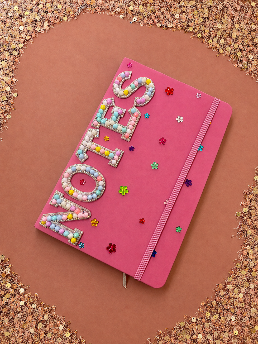 A pink notebook decorated with multicolored bead and rhinestone embellishments spelling 'LOVE' and surrounded by small rainbow-colored metallic flower confetti. The notebook has a pink elastic band closing it.