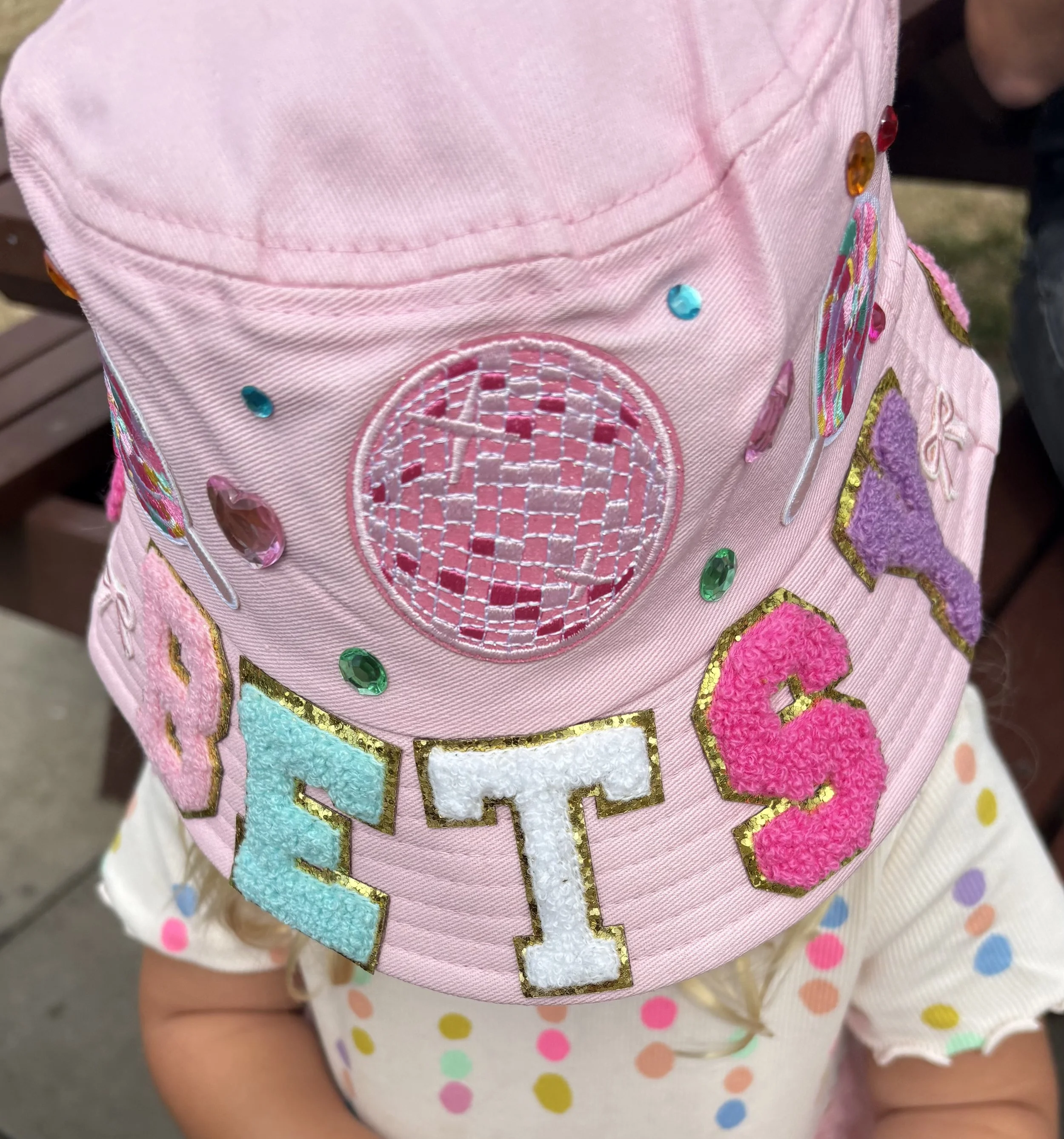 Close-up of pink fabric with colorful embroidered and beaded patches, including a globe and letter 'H', worn on a child's outfit.