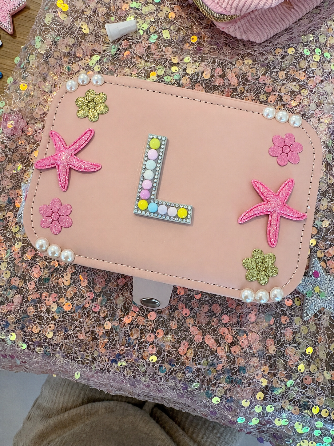 Decorative pink jewelry box decorated with starfish, flower, and letter 'L' embellishments, placed on a sequin-covered surface.