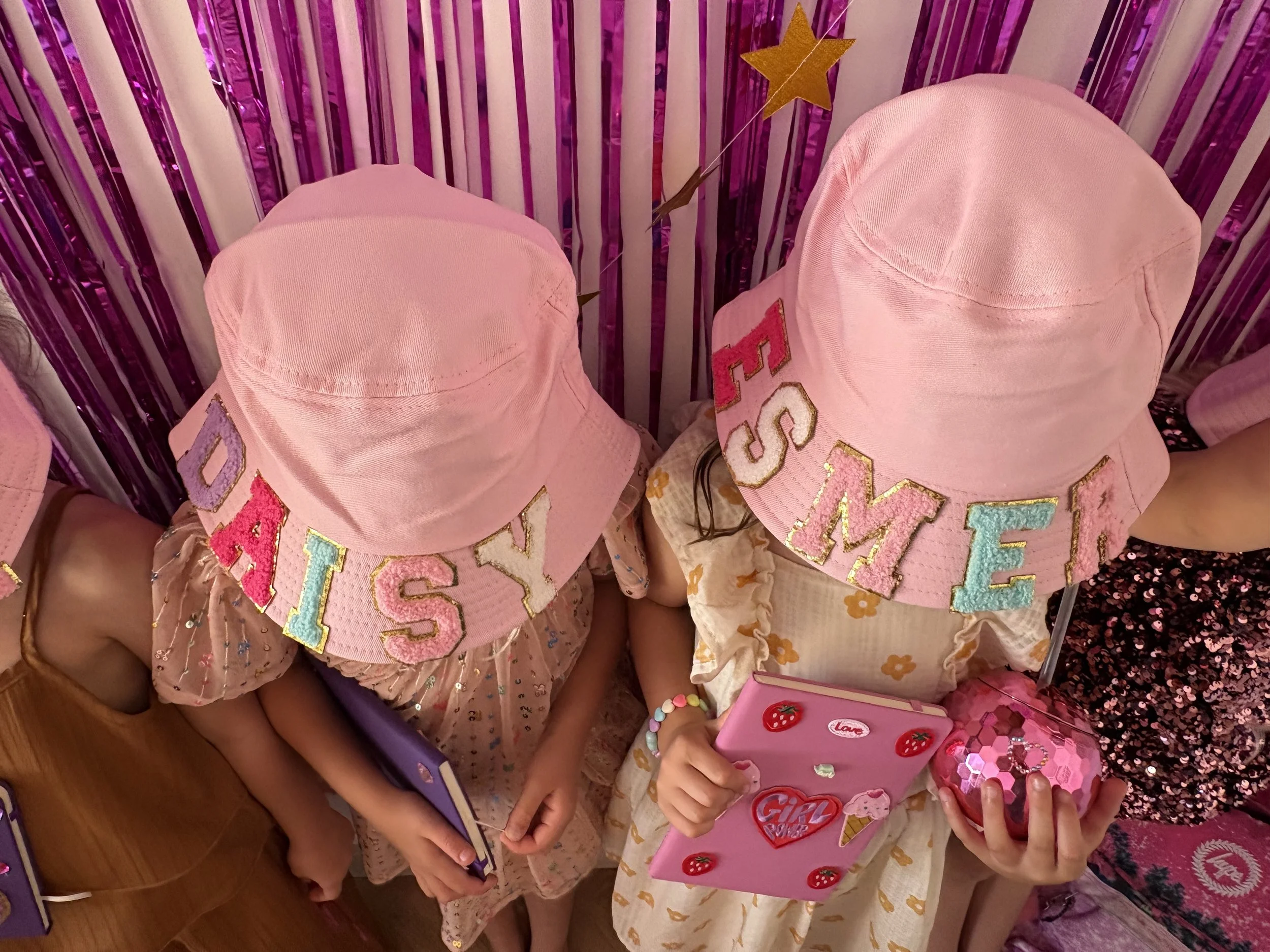 Children wearing pink hats with colorful letters, at a birthday celebration with pink decorations and a gift box with strawberries and hearts.