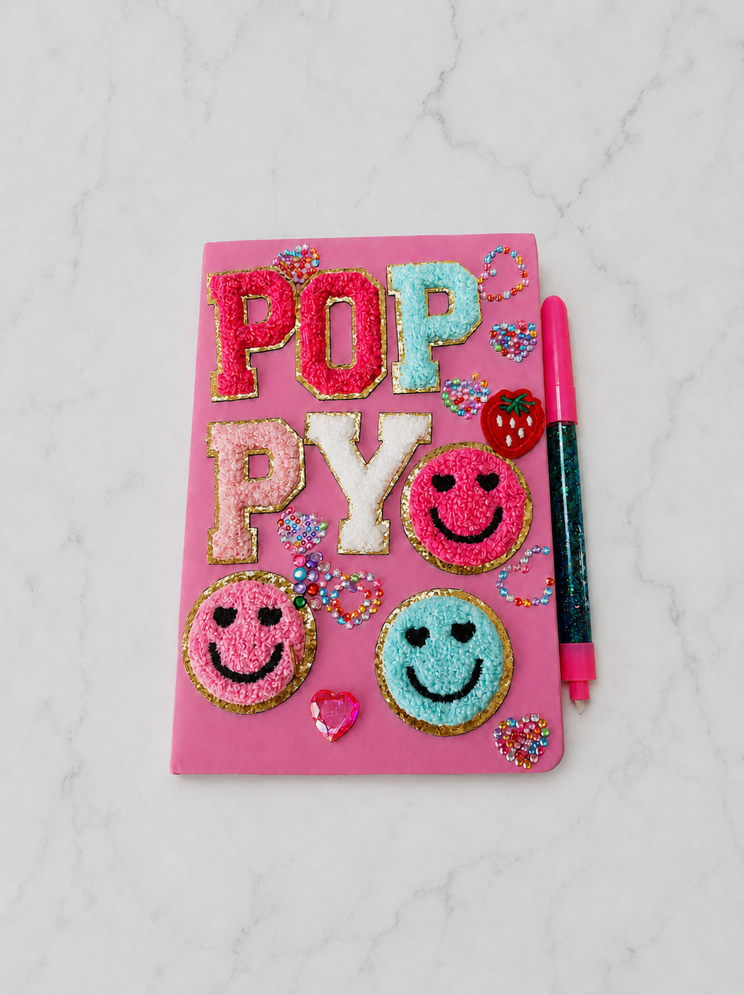 Pink notebook with decorated sugar cookies spelling out "POP PYO" and smiley faces, surrounded by colorful heart-shaped sprinkles, a strawberry-shaped pin, and a pink glitter gel pen, on a white marble surface.