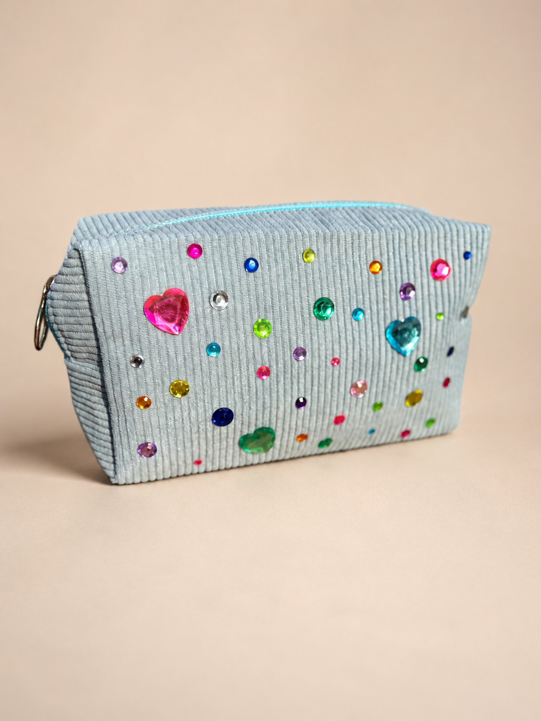 A small, light blue, corduroy purse decorated with colorful rhinestones and heart-shaped gems, placed on a beige surface.