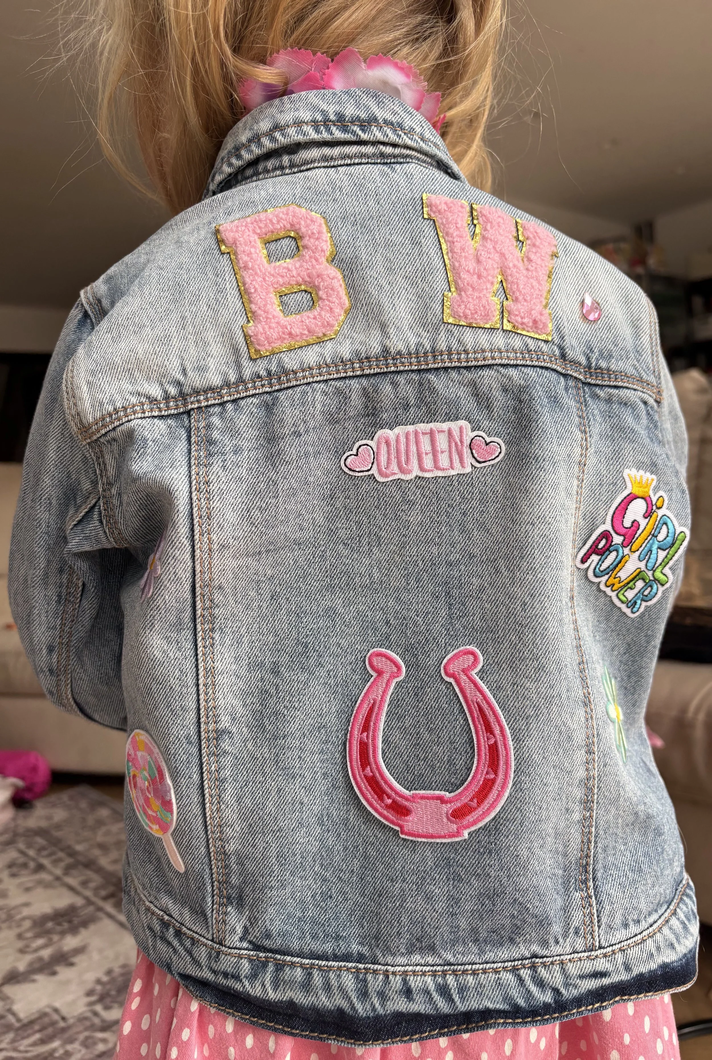 Back of a girl wearing a denim jacket with pink patches and embroidery, including the words 'B.W.', 'Queen', and decorative patches such as a horseshoe, lollipop, and other colorful designs, with a pink polka dot skirt and pink flower hair accessory.