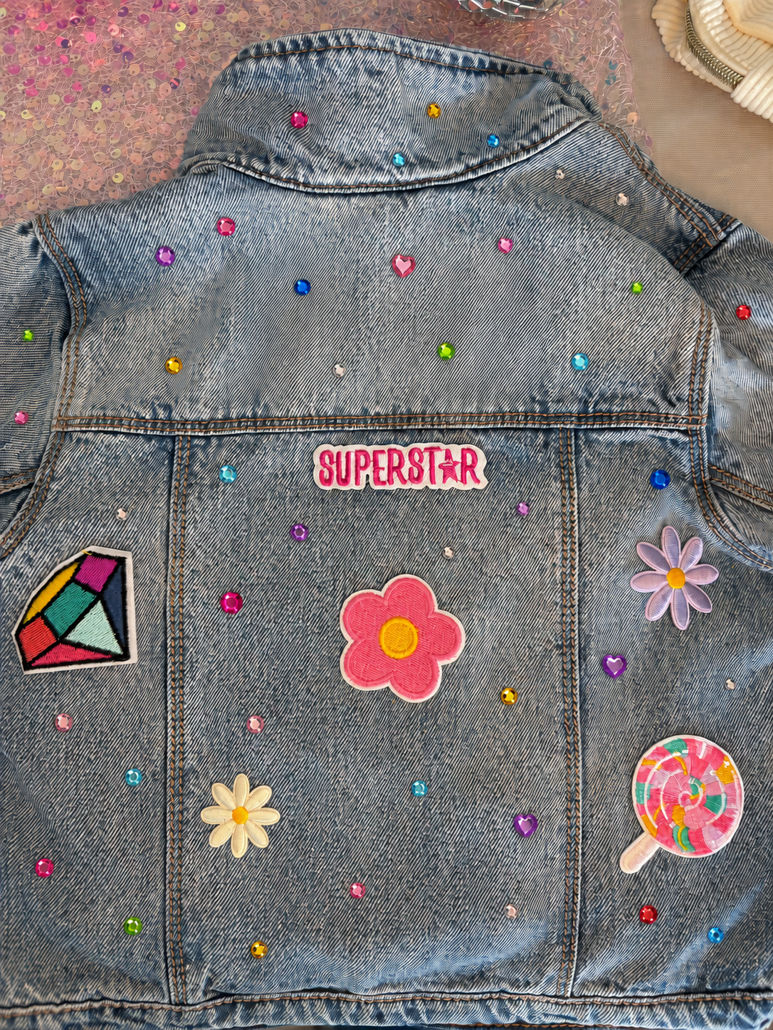 Back of a denim jacket decorated with colorful patches, rhinestones, and stickers, including a pink flower, a daisy, a rainbow diamond, a pink lollipop, and the word 'SUPERSTAR'.