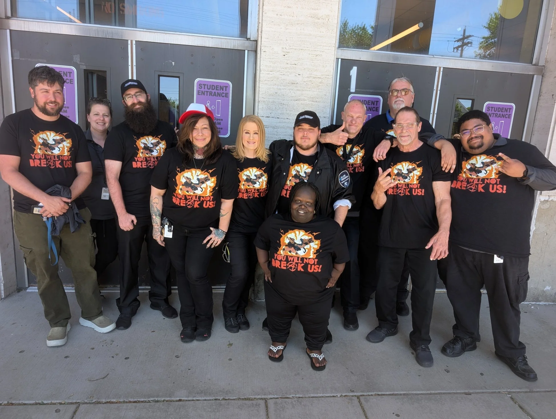 A group of twelve people standing outside a building with purple 'Student Entrance' signs on the doors, wearing matching black T-shirts with a graphic and the text 'You Will Not Break Us!'