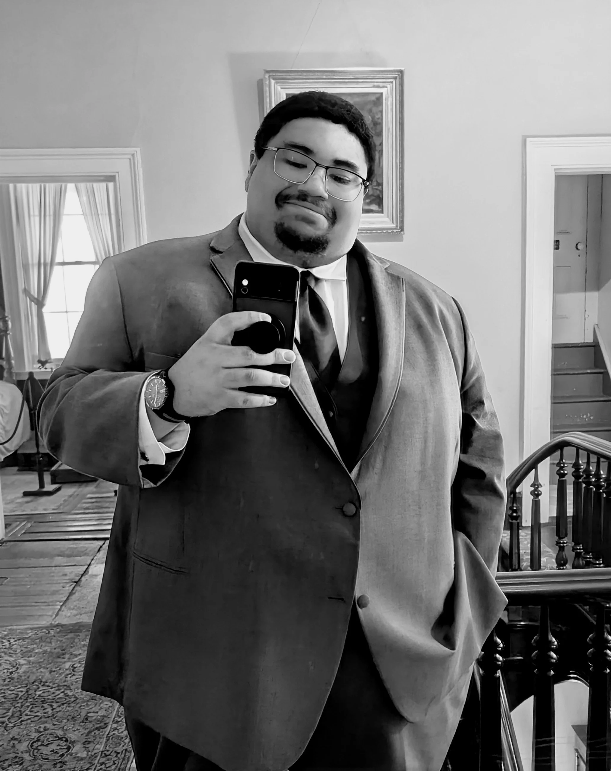 A man wearing glasses and a suit is taking a selfie in a mirror with a smartphone. The setting appears to be a home or a room with a staircase, paintings, and large windows with curtains.