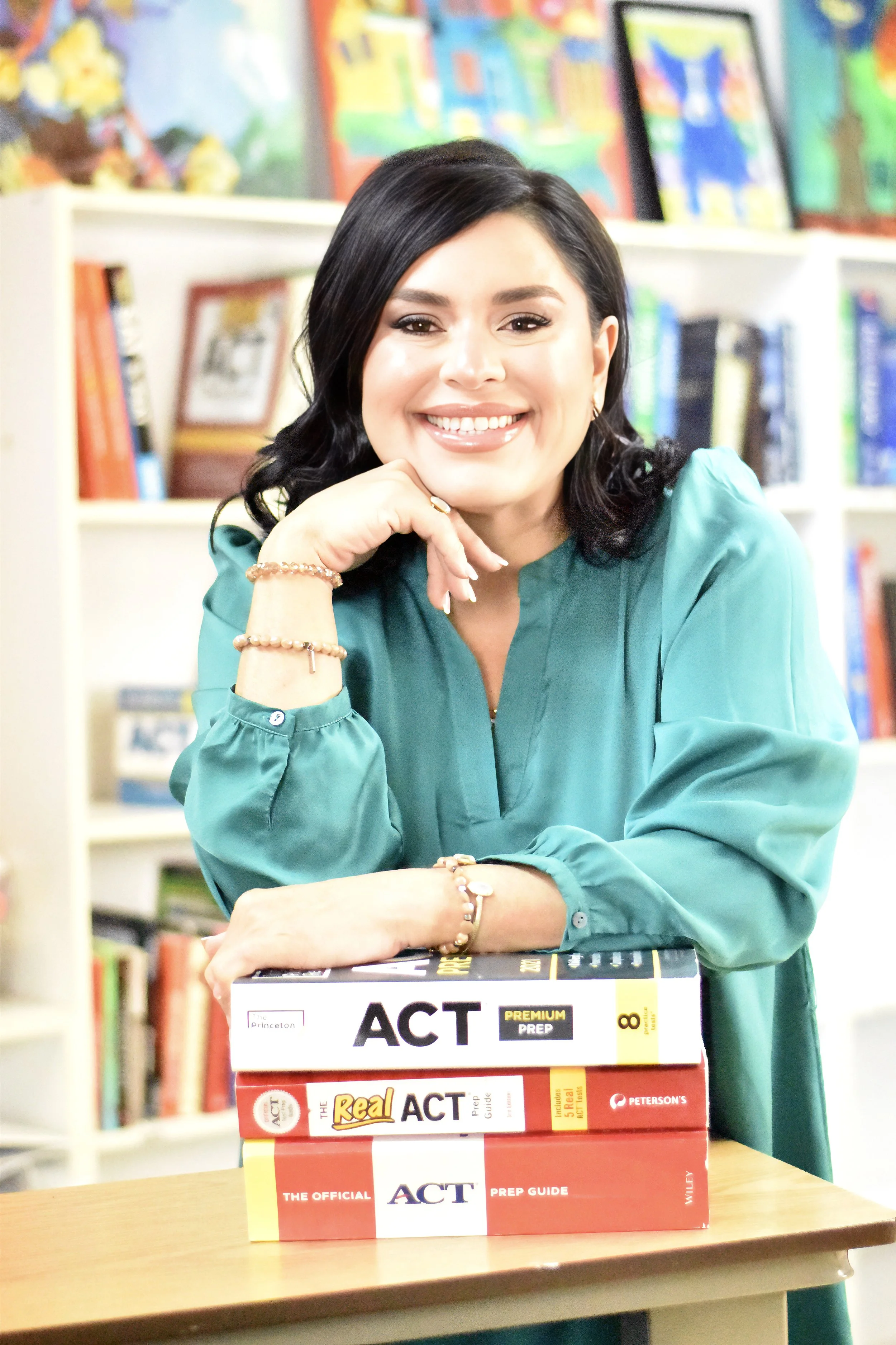 A woman with black hair and a teal blouse sitting in a library or classroom, smiling at the camera. She is resting her chin on her right hand, and on the table in front of her are multiple ACT prep books.