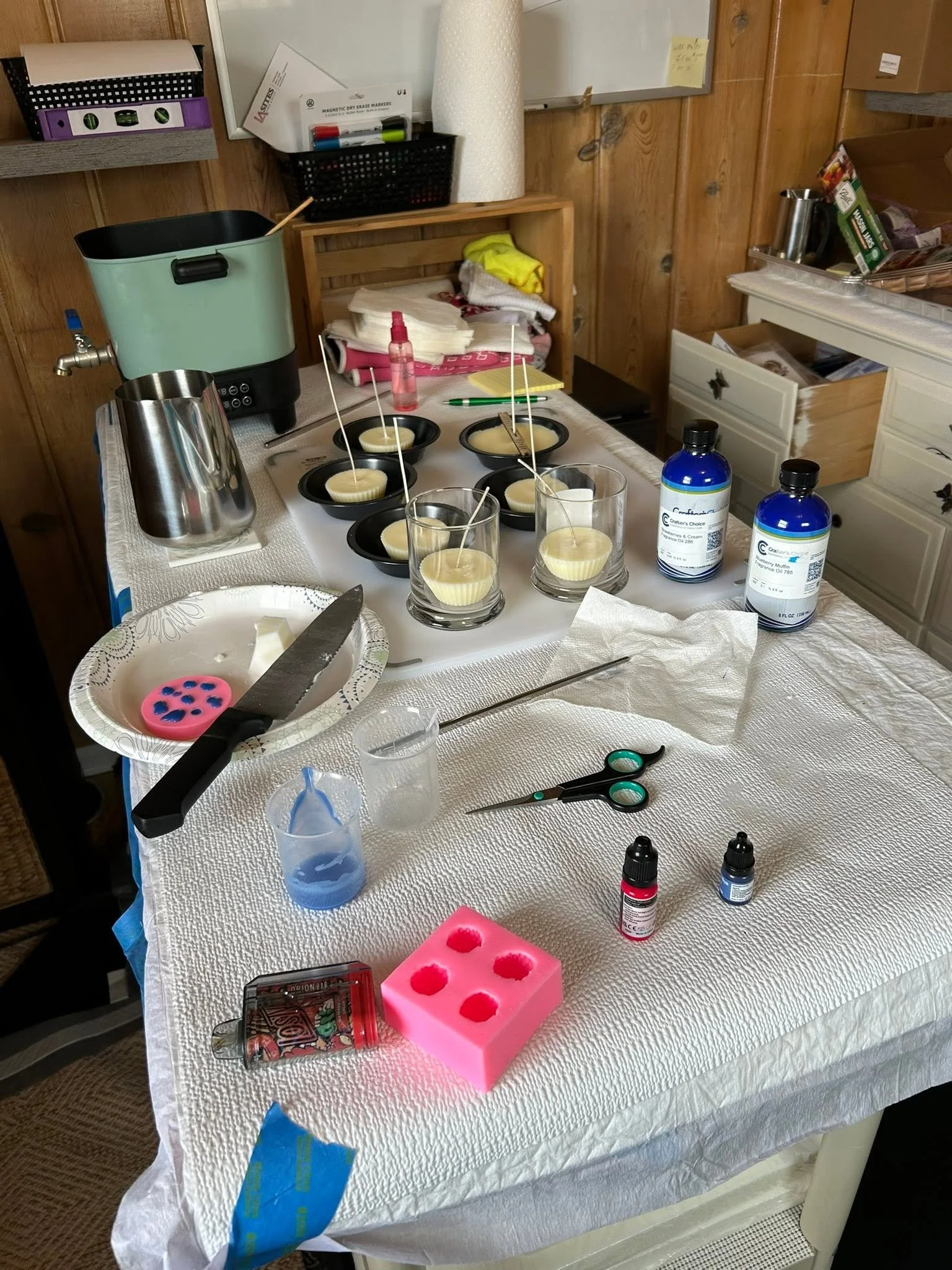 A kitchen or craft room table with supplies including silicone muffin molds filled with a white waxy substance, a pink silicone mold, various bottles, scissors, a knife, and other miscellaneous items. The background has wood paneling, storage boxes, a paper towel roll, and a whiteboard with notes.