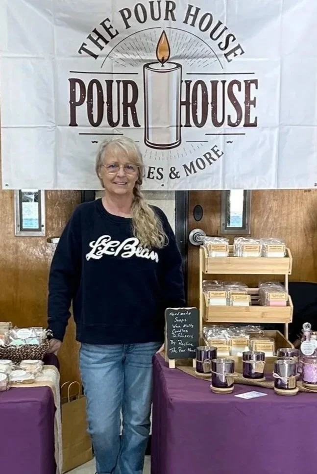 A woman standing behind a table with soap products at a booth labeled 'The Pour House.' The banner behind her features a candle and the words 'The Pour House Soap & More.'