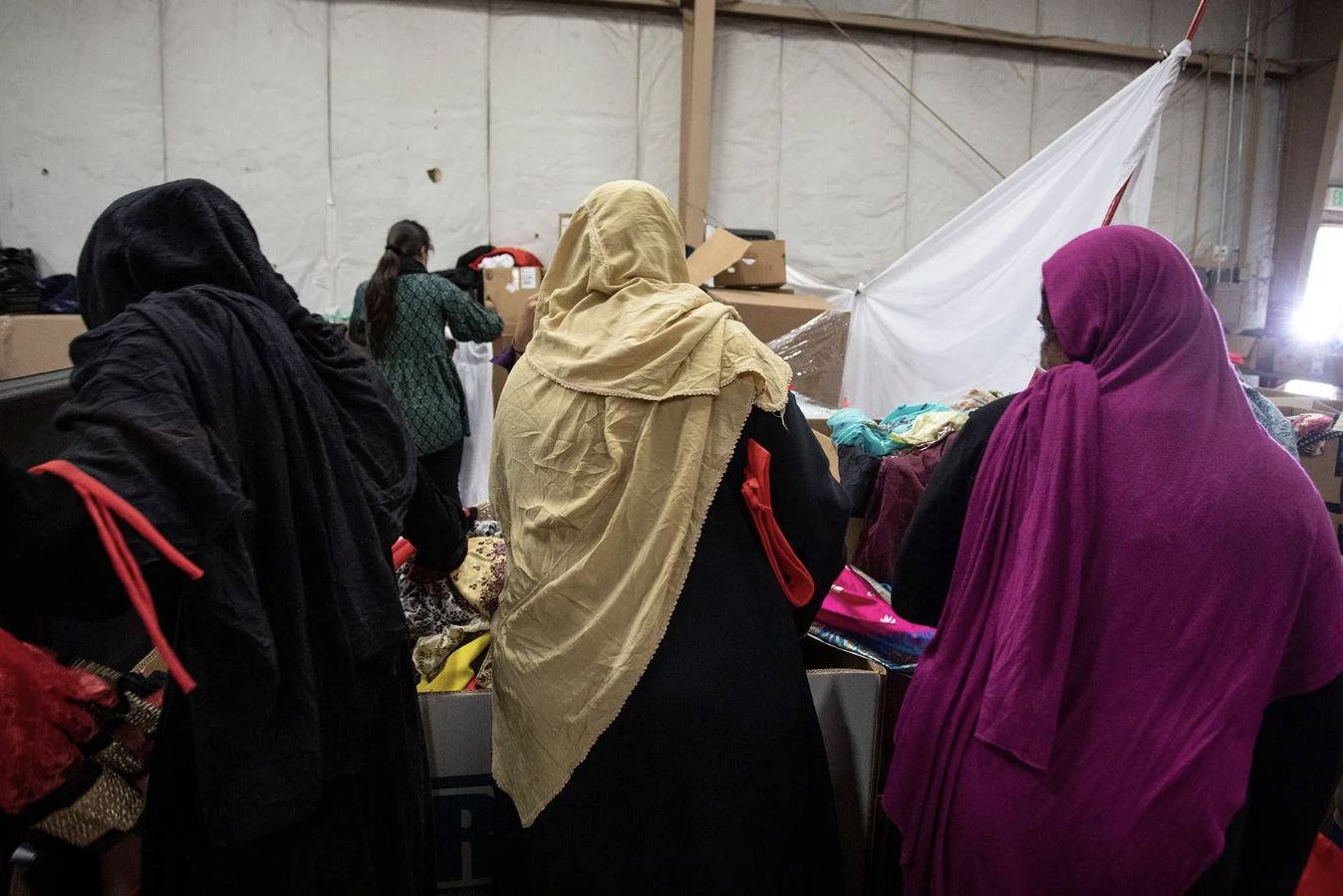 Four women wearing headscarves shopping for clothes at an indoor market or thrift store, with various items on tables and boxes around them.