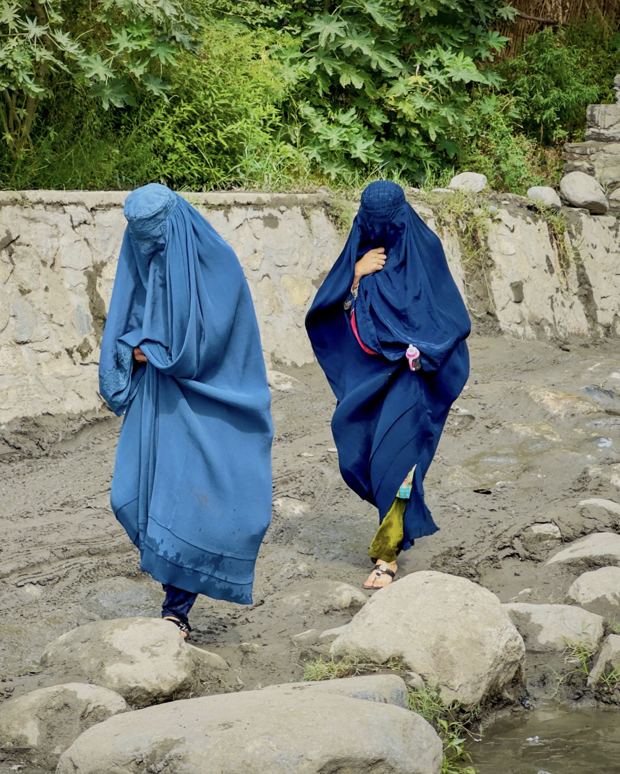 Two women dressed in blue traditional clothing walking on a rocky dirt path near a stone wall and green foliage.