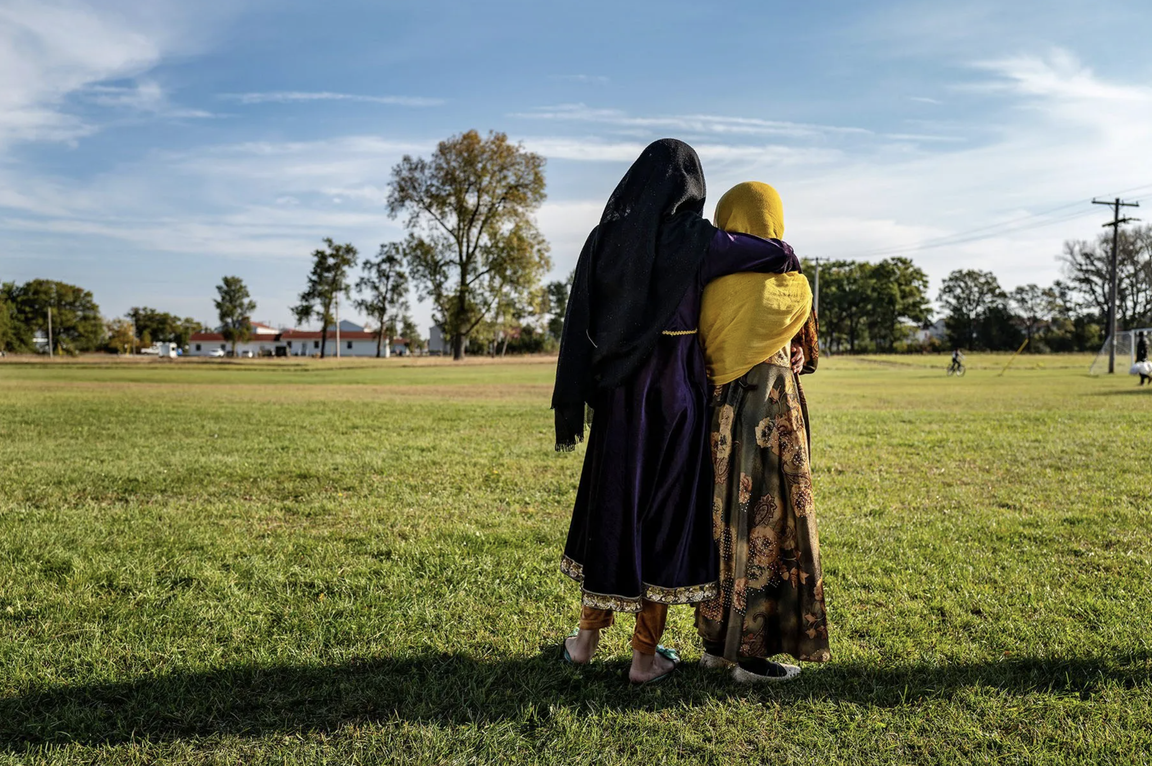 Two women dressed in traditional attire hugging on a grassy field on a sunny day, with trees, houses, and power lines in the background.