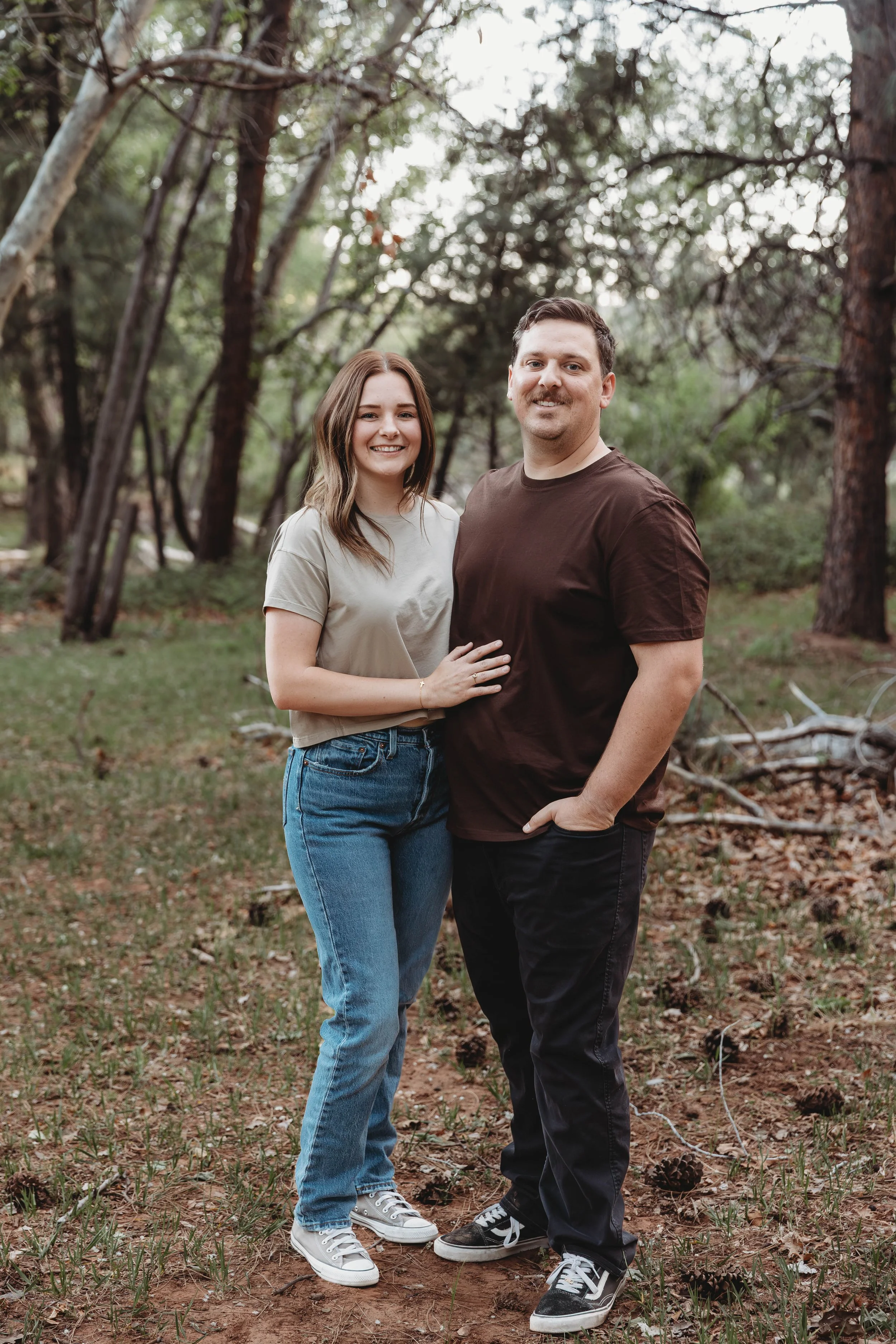 A smiling young woman with brown hair, wearing a beige t-shirt, jeans, and sneakers, standing next to a smiling man with short dark hair, wearing a dark brown t-shirt, black pants, and sneakers, outdoors in a wooded area with trees and green grass.