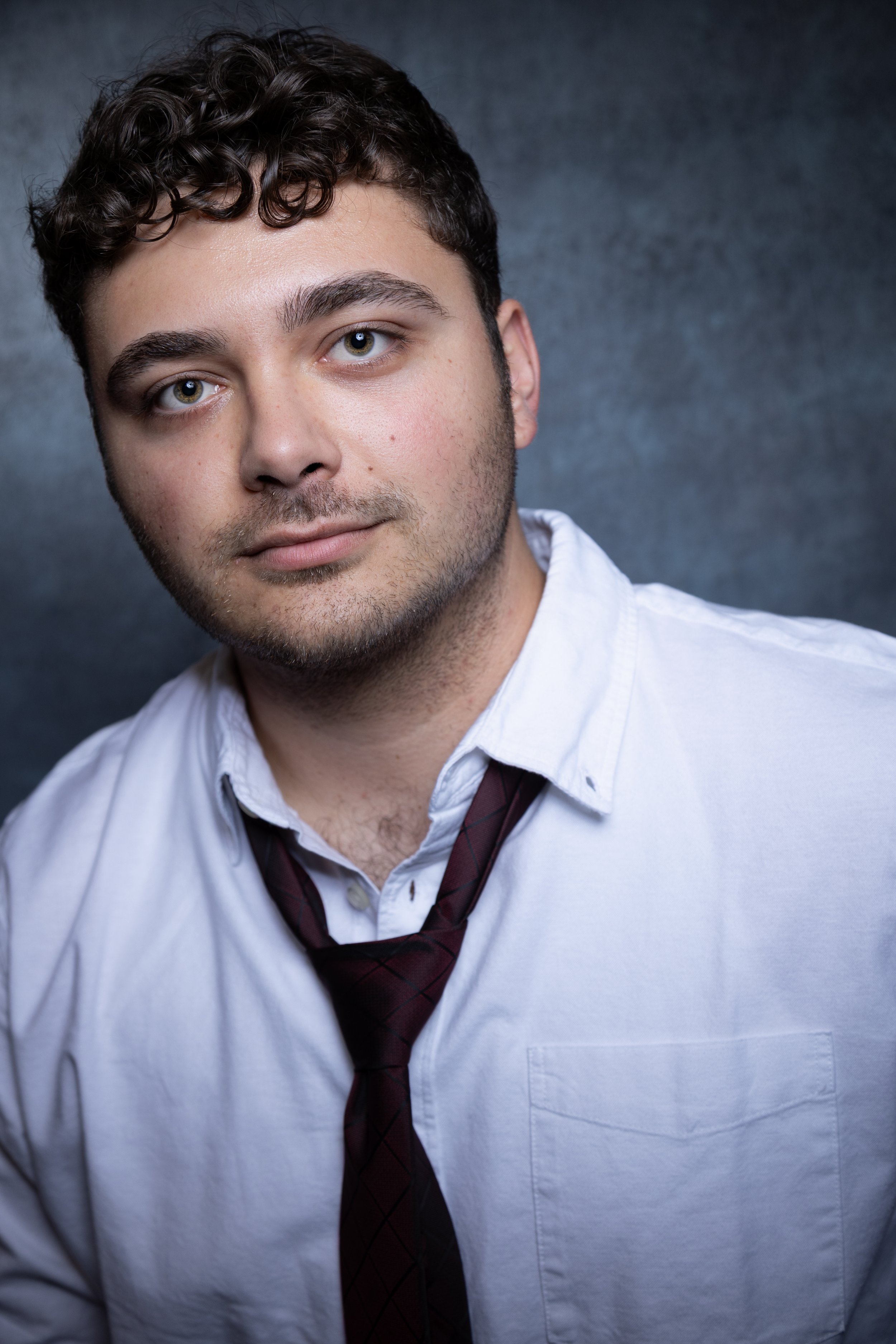 A young man with curly brown hair, hazel eyes, and light skin, wearing a white dress shirt and a maroon tie with black diamond pattern, against a dark background.