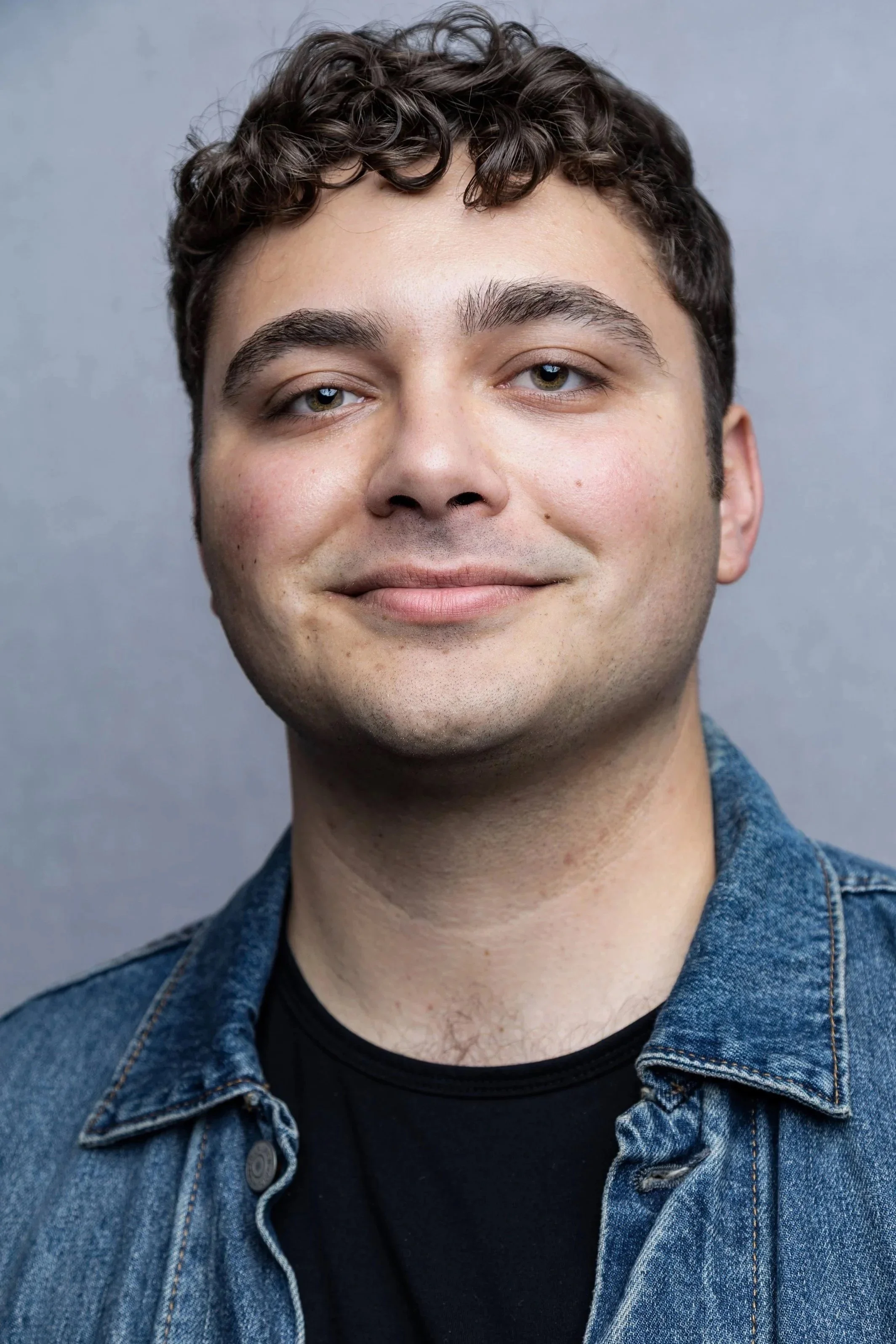 Close-up of a young man with curly dark hair and light-colored eyes, wearing a denim jacket over a black shirt, smiling softly against a gray background.