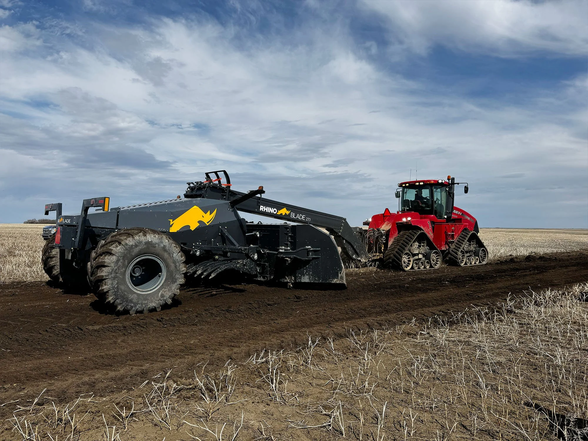 A large red tractor pulling a black agricultural implement through a harvested field with dry stalks; the sky is partly cloudy.