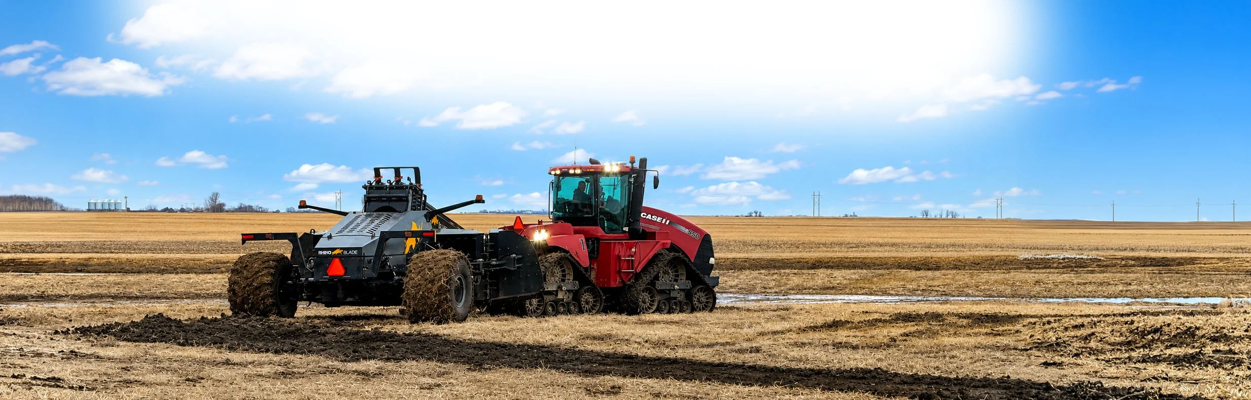 A red Case IH tractor pulling a black Rhino Blade in a large farm field under a blue sky with scattered clouds.