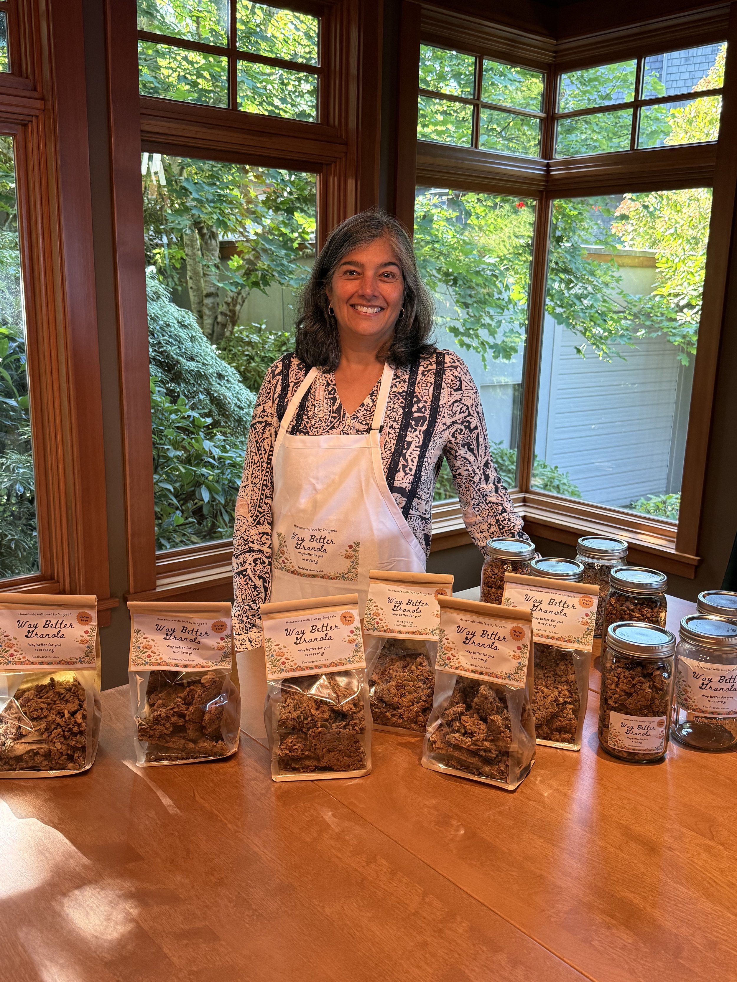 Smiling woman standing behind a table with various jars and bags of granola labeled 'Way Better Granola,' inside a room with large windows showing a green outdoor garden.
