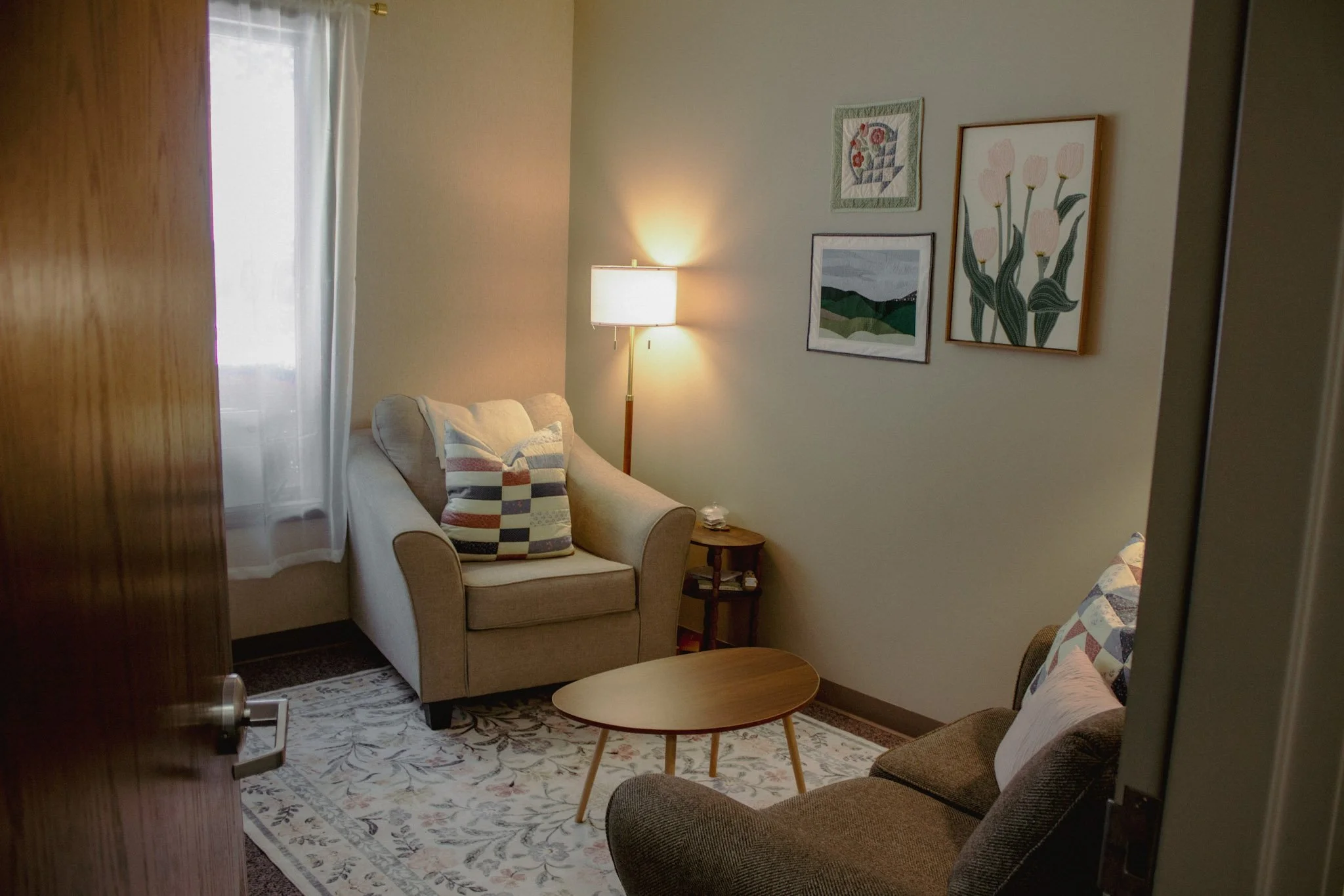 Cozy living room with beige sofa, patterned pillows, a wooden side table, a floor lamp, framed floral and landscape artwork on the wall, and a patterned rug.