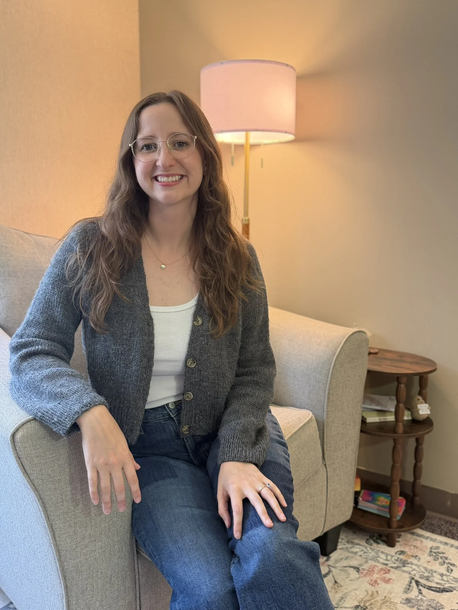 A woman with wavy brown hair, wearing glasses, a white top, and a gray jacket, smiling while sitting on a beige armchair in a cozy living room with a floor lamp and a side table.