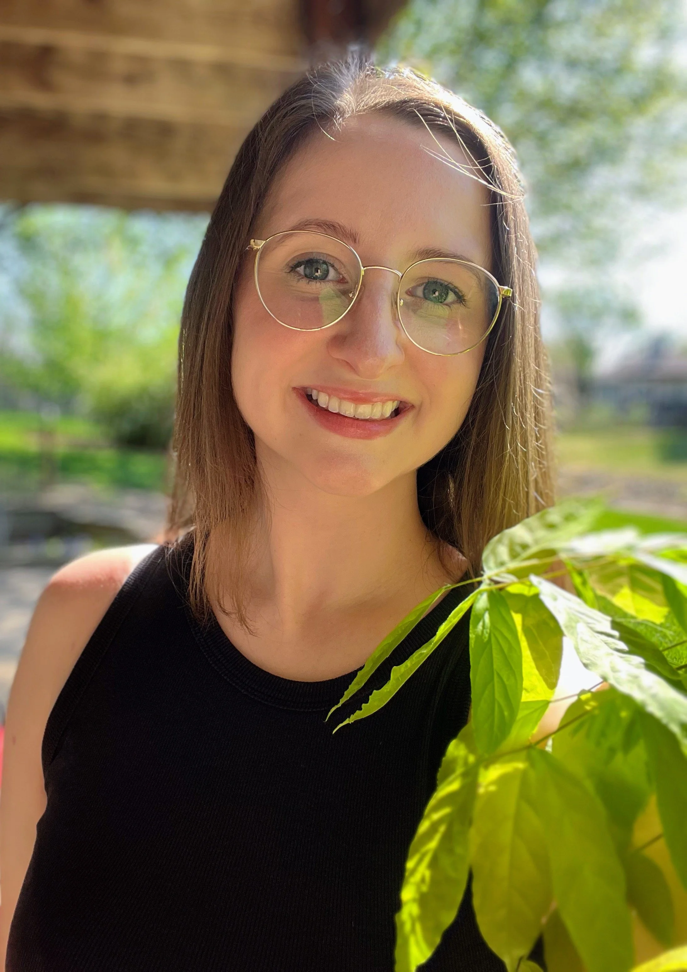 Close-up of a young woman with light brown hair, wearing round glasses and a black sleeveless top, smiling outdoors with green trees in the background and sunlight illuminating her face.