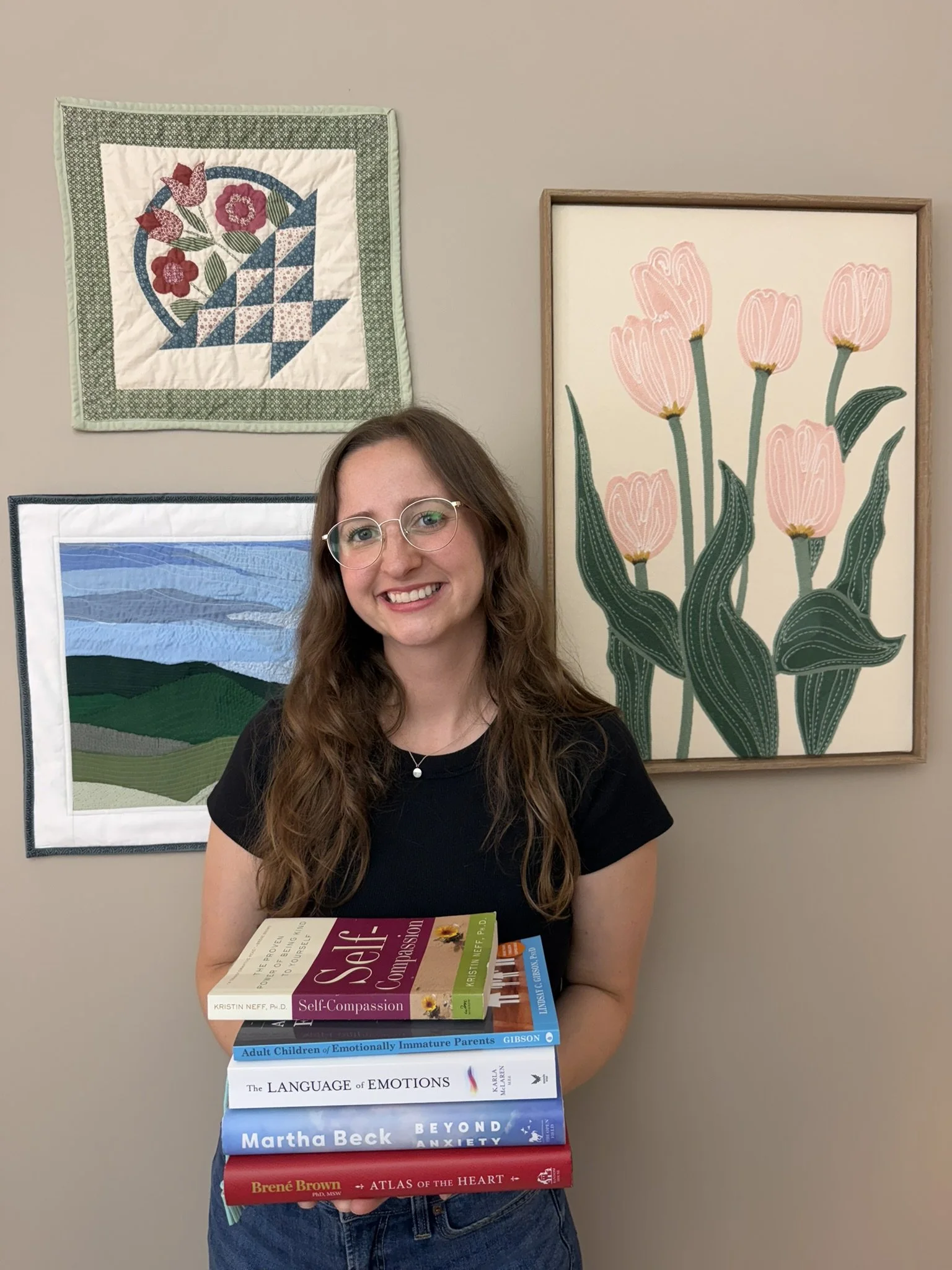 A young woman with long wavy hair and glasses smiling and holding a stack of books in front of her, standing in front of a beige wall decorated with framed artwork including a quilt and floral paintings.