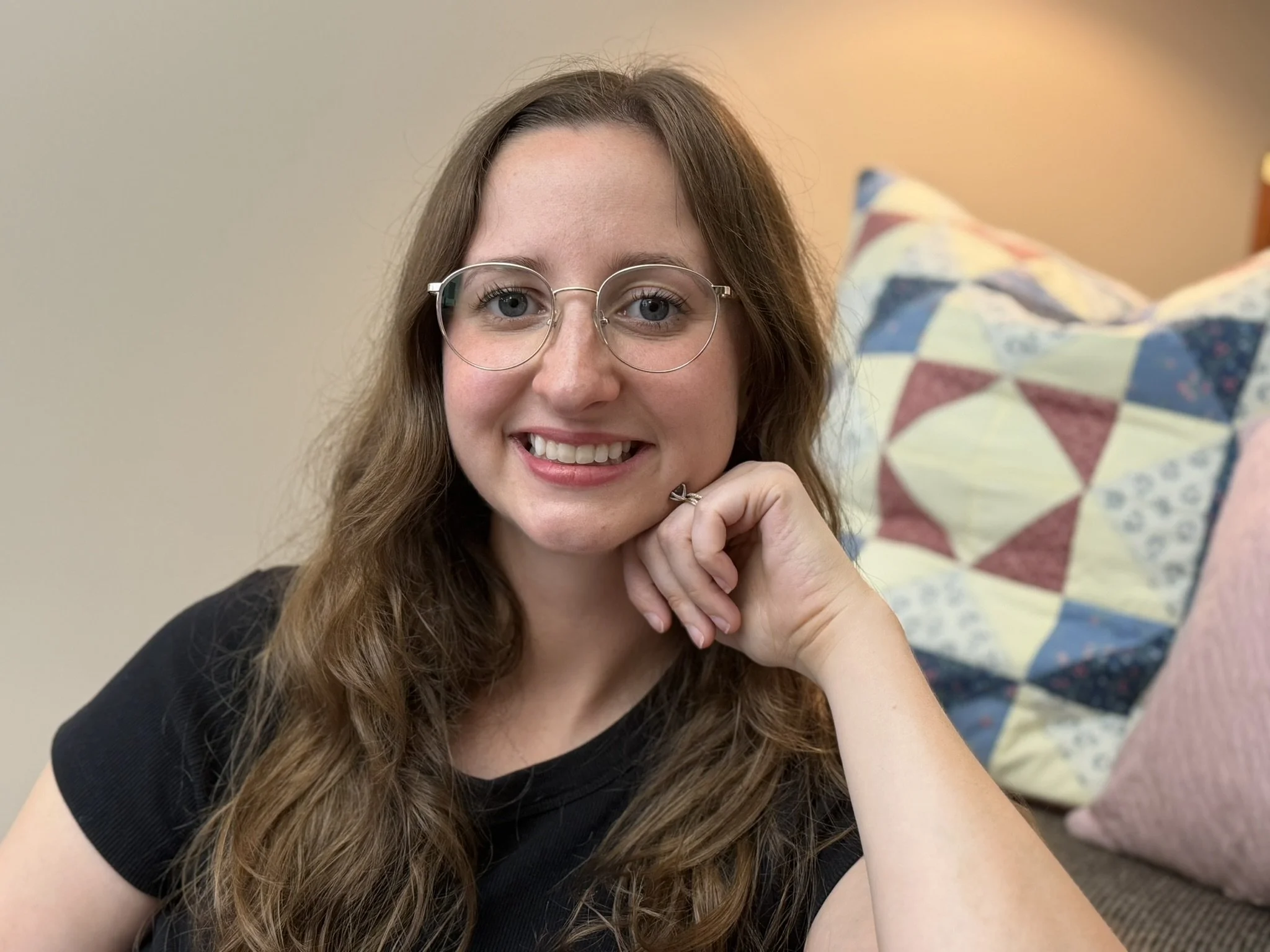 A young woman with long curly brown hair and glasses smiling at the camera, resting her chin on her hand, with a pillow with a colorful patchwork quilt pattern in the background.