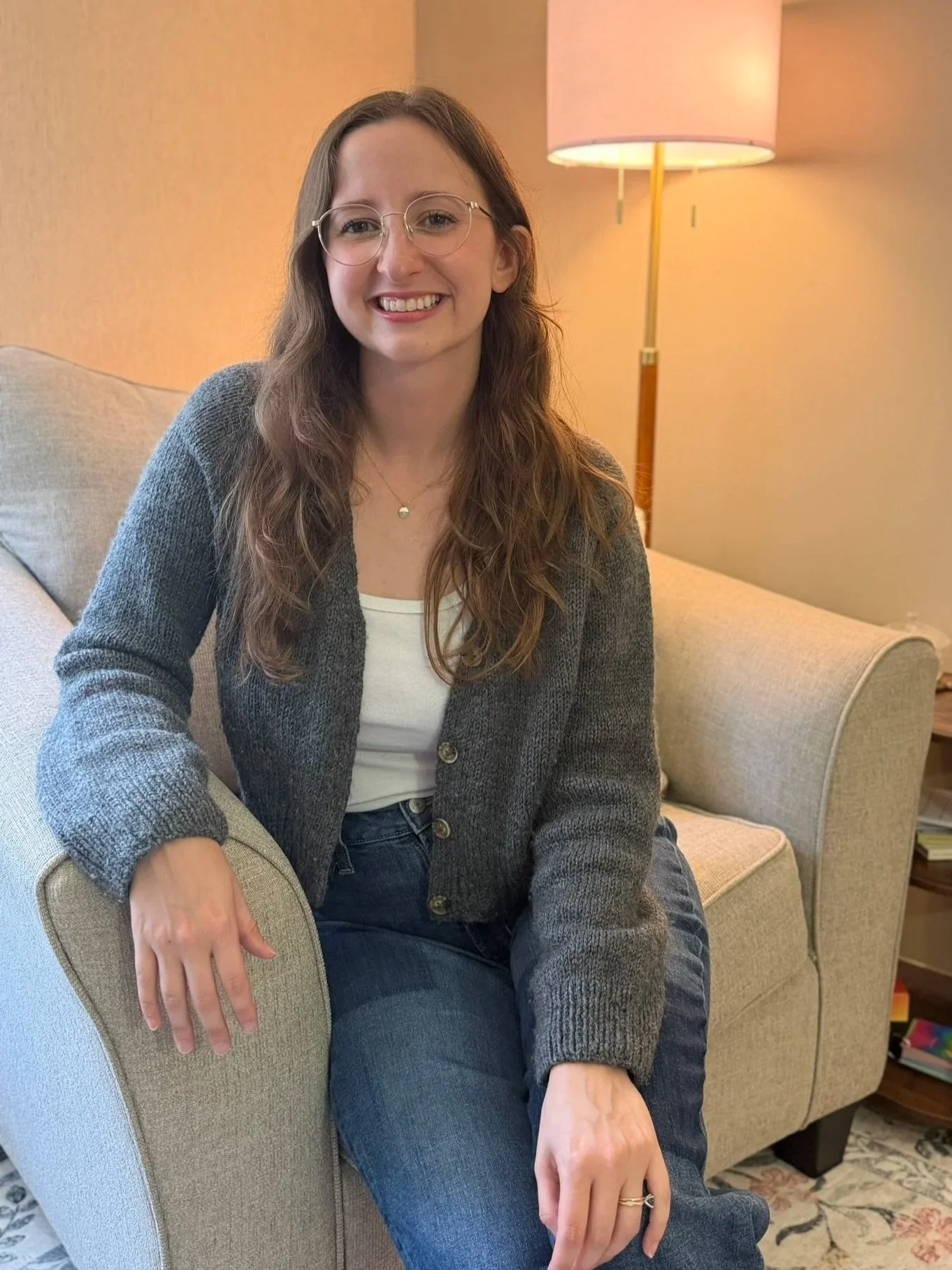 A smiling woman with glasses and long brown hair sitting on a beige armchair in a cozy living room with a floor lamp and a patterned rug.