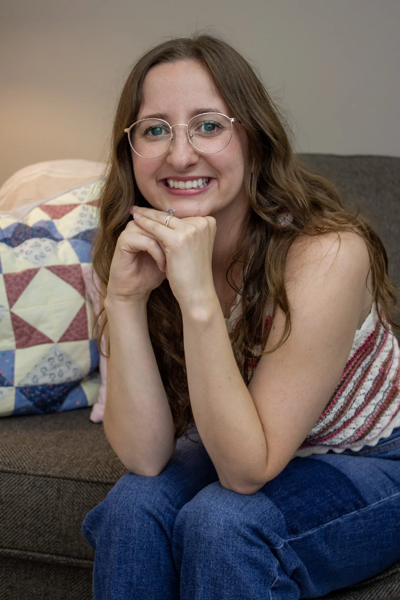 A woman with long brown hair, glasses, and a bright smile sitting on a couch, with her hand under her chin, wearing a striped top and blue jeans, in a cozy living room with a quilt on the backrest.
