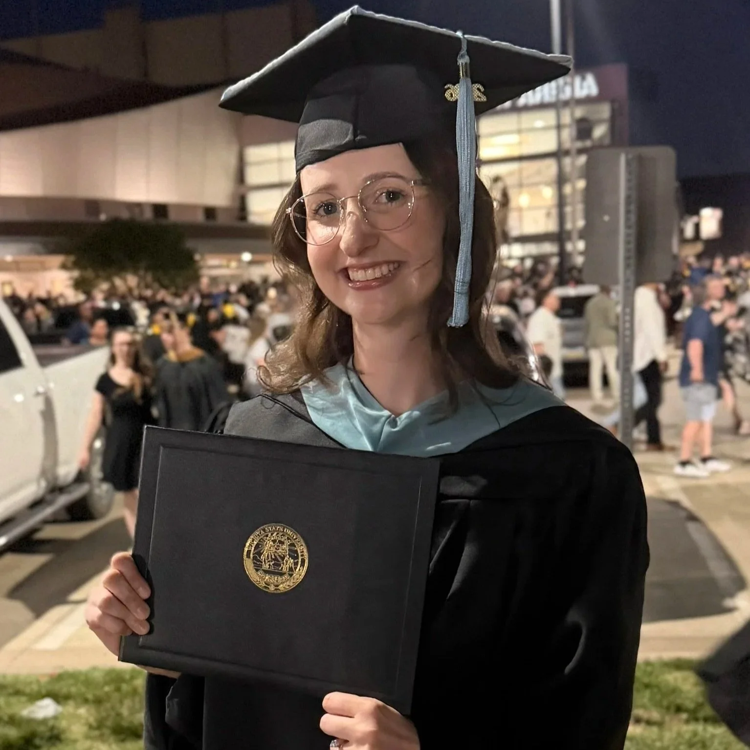 A woman dressed in graduation cap and gown holding a diploma in front of a crowd at an outdoor graduation ceremony at night.