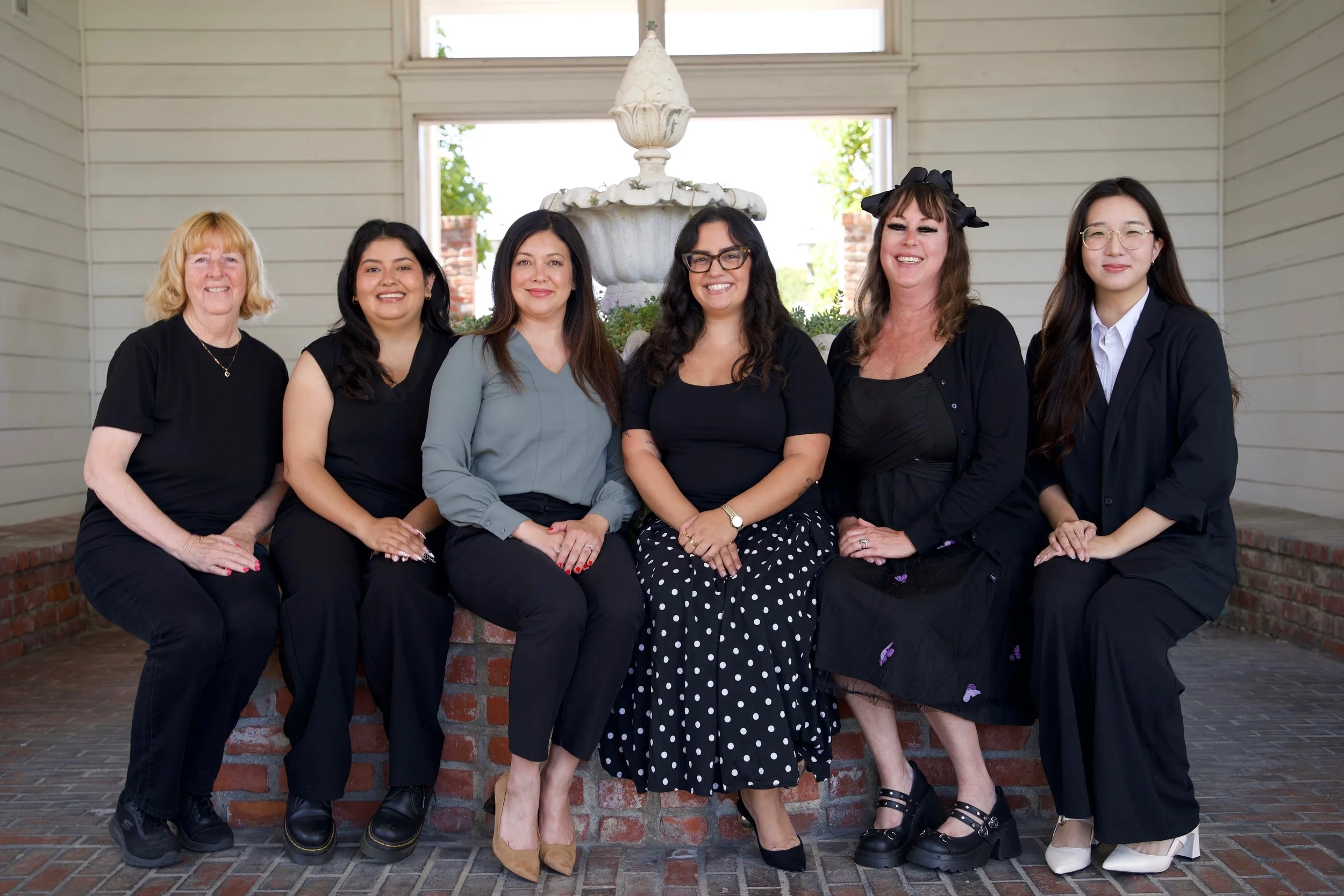 Group of six women sitting on a brick bench outside a house with white wooden walls and a fountain in the background.