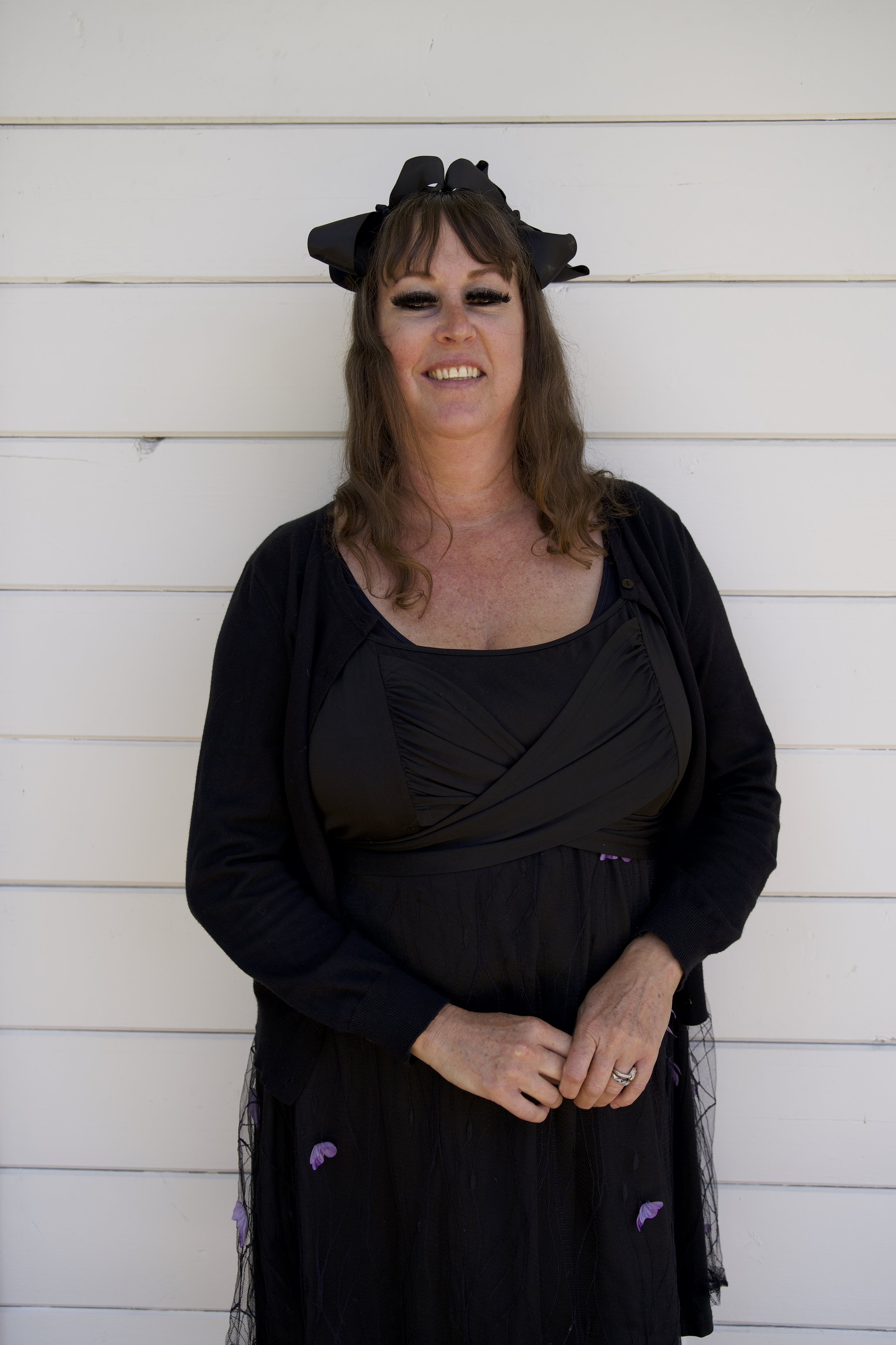 A woman with brown hair, wearing a large black bow headband, black makeup, a black dress, and a black cardigan, standing in front of a white horizontal panel wall.