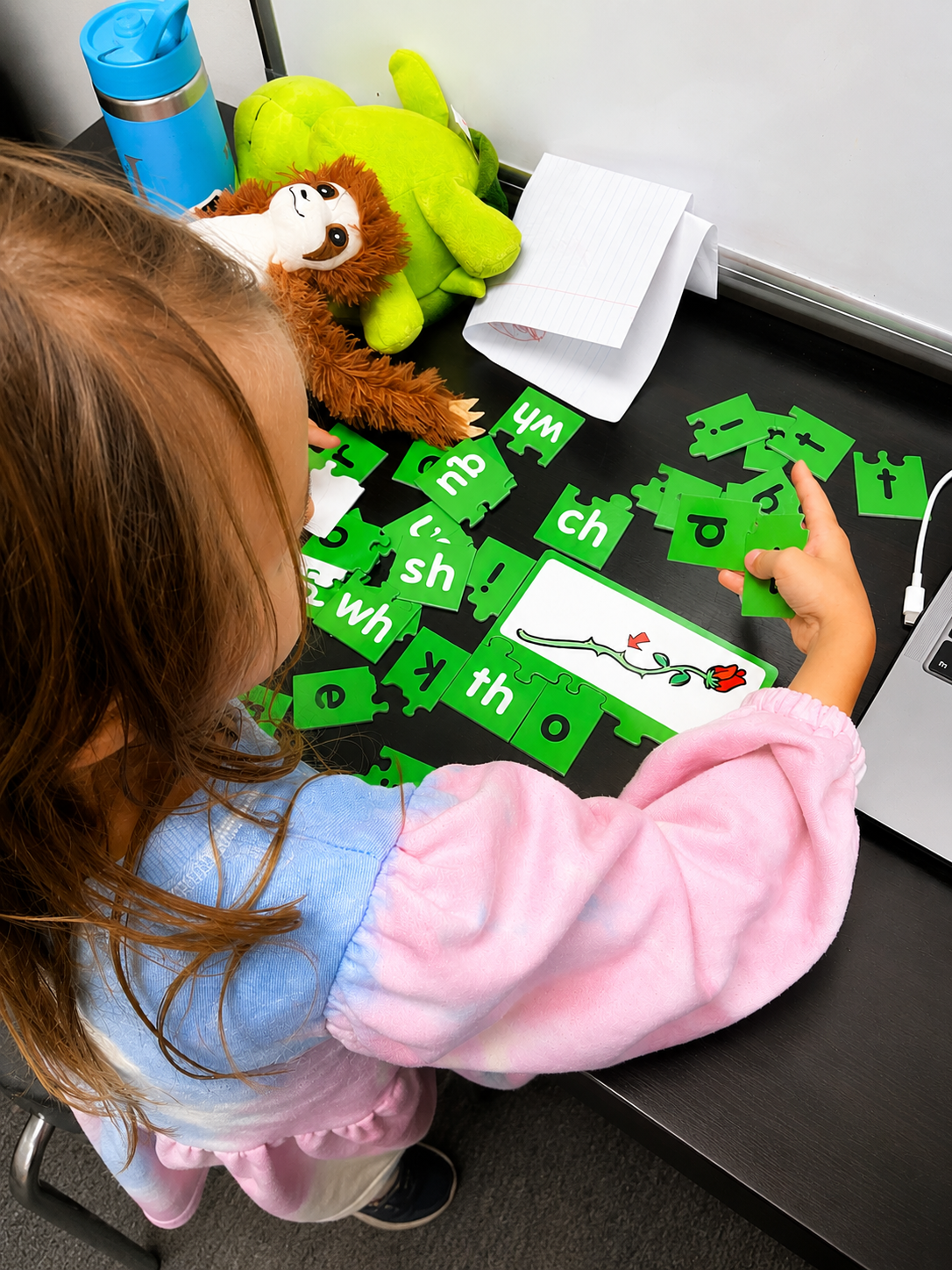 Child wearing a pink and blue long sleeve shirt playing with green puzzle pieces and a picture of a flower on a puzzle card, surrounded by plush toys including a lion and a turtle, with a blue water bottle and a notebook nearby.
