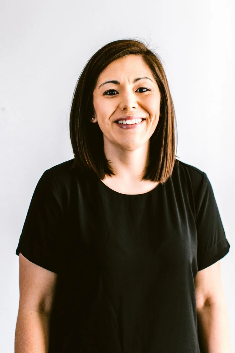 A smiling woman with shoulder-length brown hair wearing a black T-shirt standing against a plain white wall.