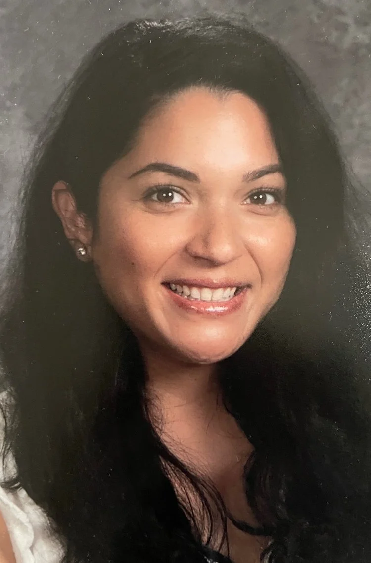 Portrait of a young woman with long black hair, smiling, wearing earrings and a light-colored top.