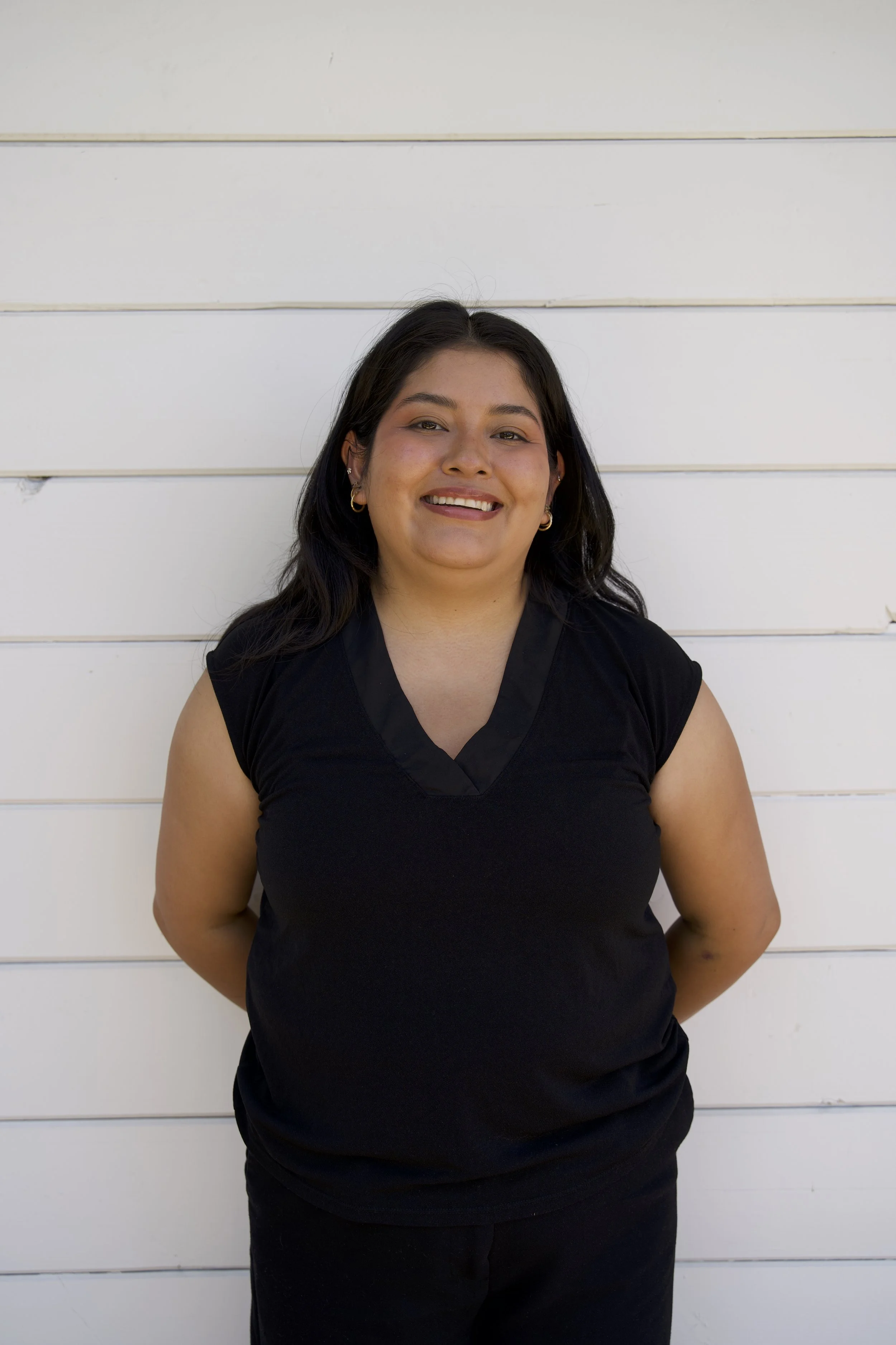 A woman with long dark hair smiling, standing against a white horizontal panel wall, wearing a black sleeveless top and gold hoop earrings.